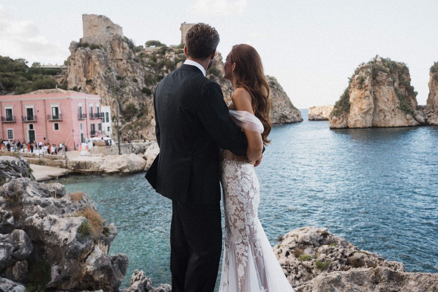 Bride and groom embracing while overlooking stunning turquoise cove with colorful buildings and dramatic rock formations.