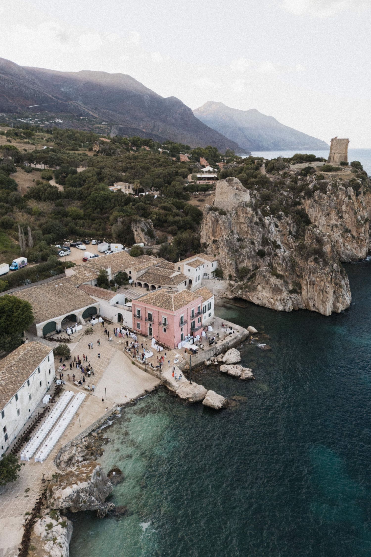 Aerial view of a coastal wedding venue with pink building nestled between cliffs and blue water.