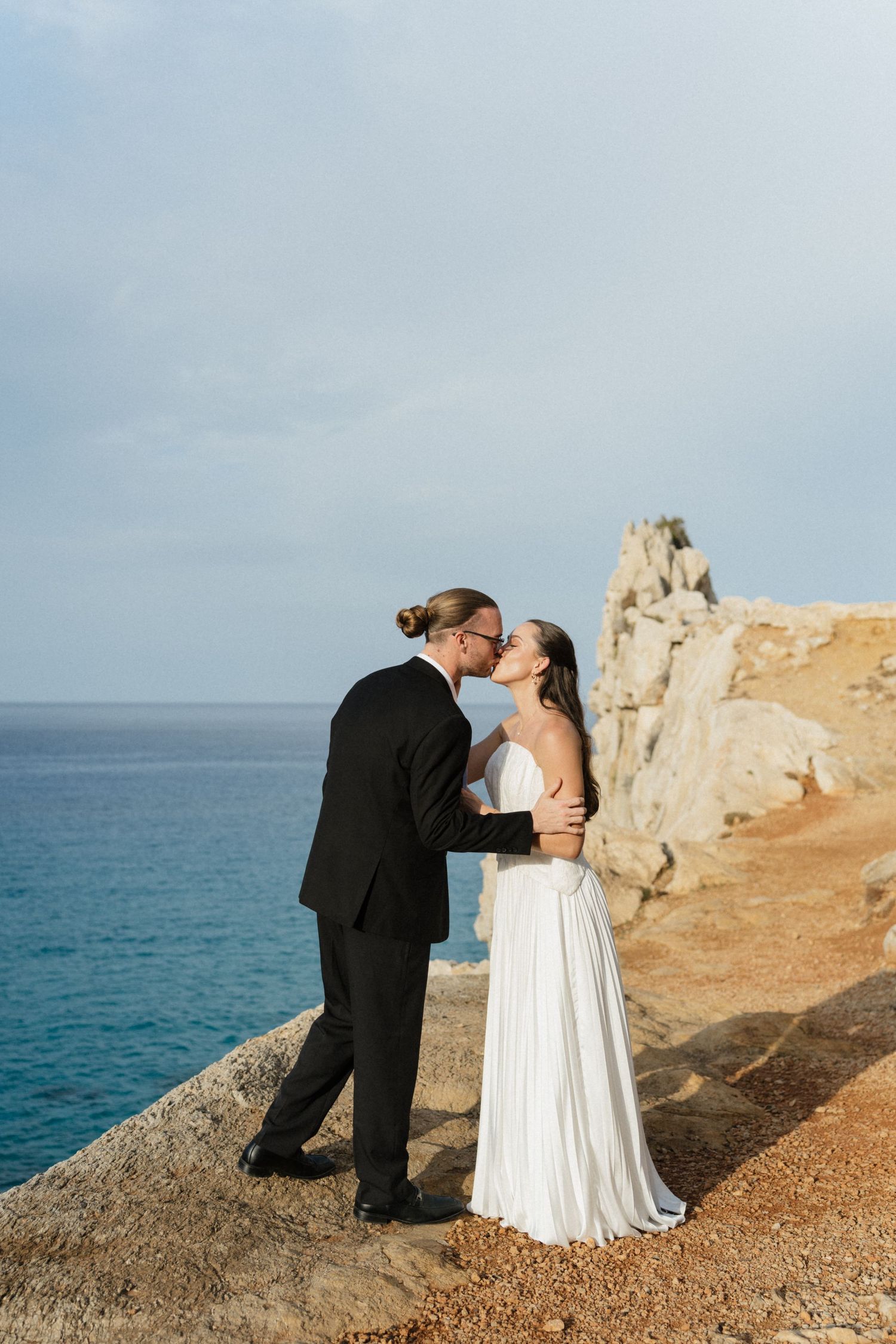 Bride in a white gown and groom in a black suit share a kiss on a rocky seaside cliff overlooking the blue sea.