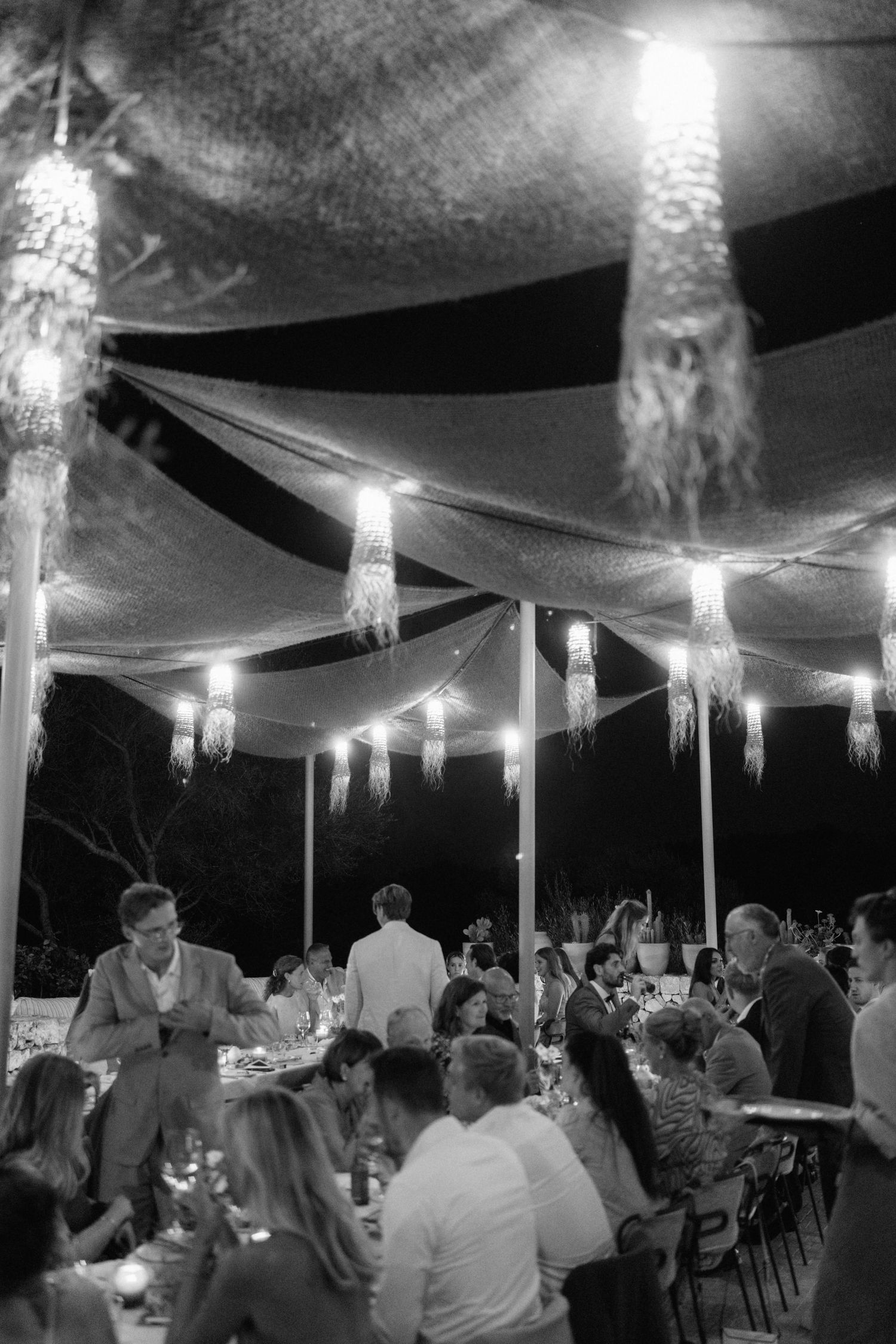 Black and white night reception under draped canopies with glowing tassel lanterns as guests dine and mingle at long tables.