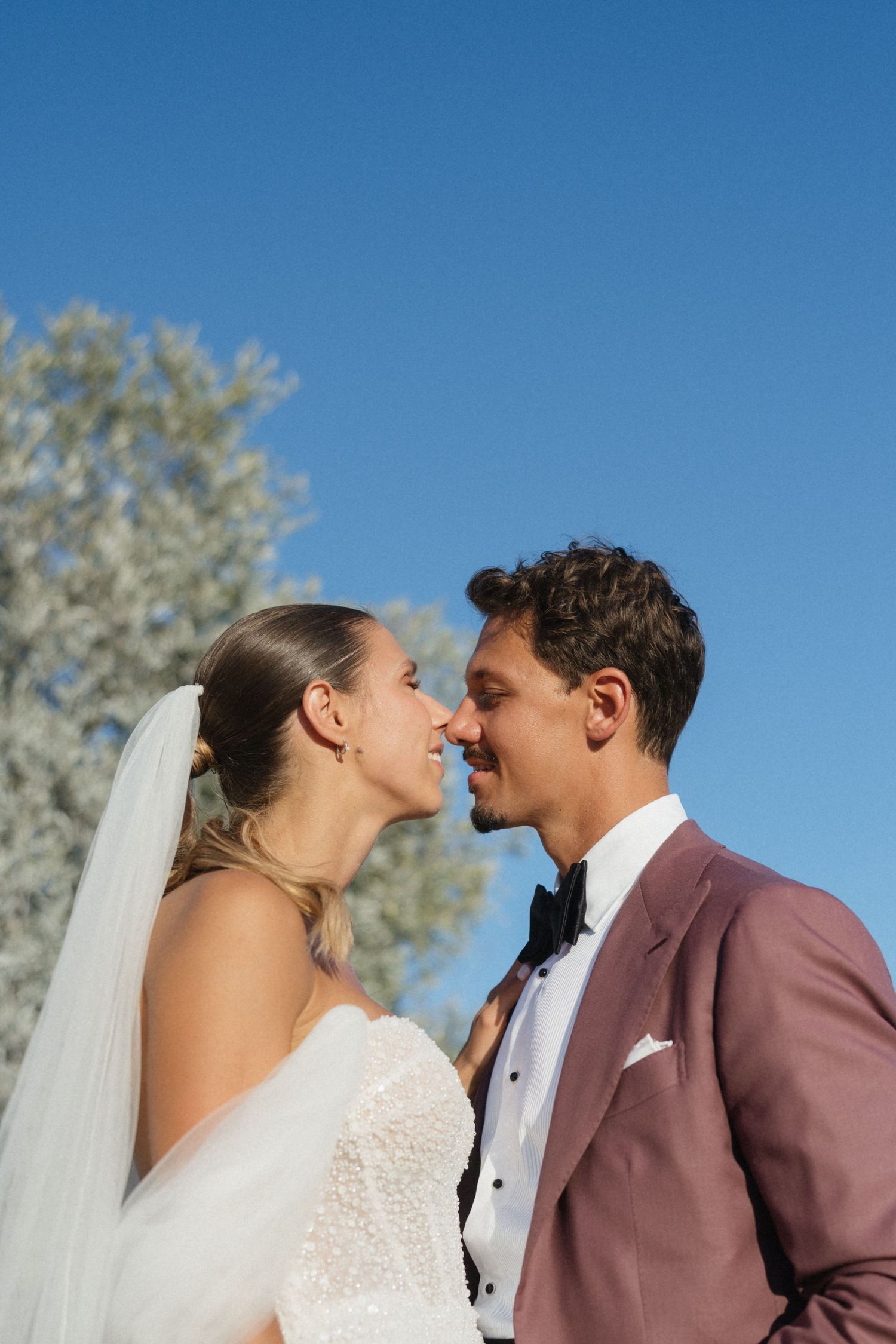 Bride in a lace gown and veil nose to nose with her groom in a mauve suit and bow tie against a clear blue sky.