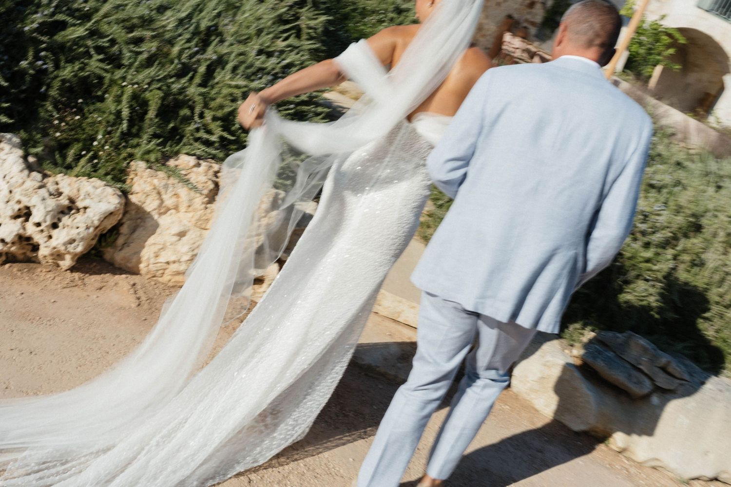Bride in a lace gown with a long flowing veil walks beside a groom in a light blue suit along a sunny Mediterranean path.