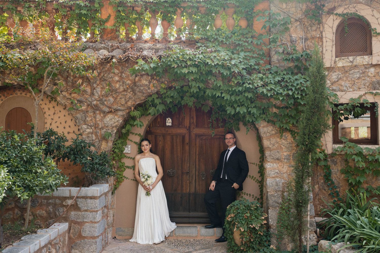 Bride in a white gown and groom in a dark suit pose before a wooden door under an ivy-covered stone archway.