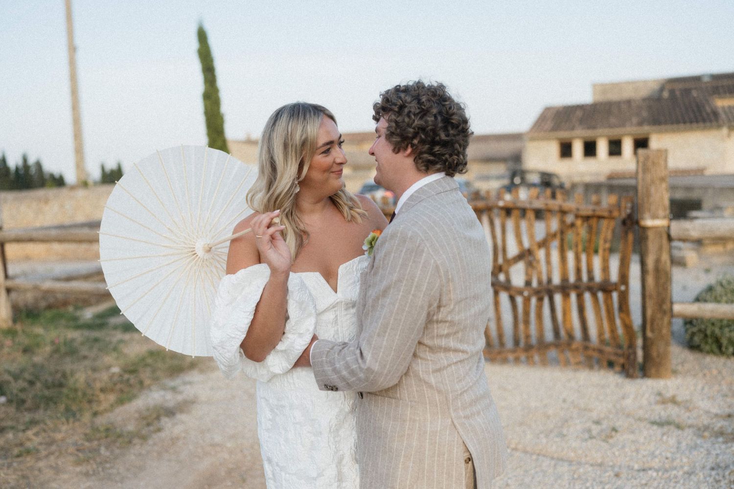 Bride and groom embracing outdoors, bride holding white parasol, groom in striped suit, rustic farm backdrop.