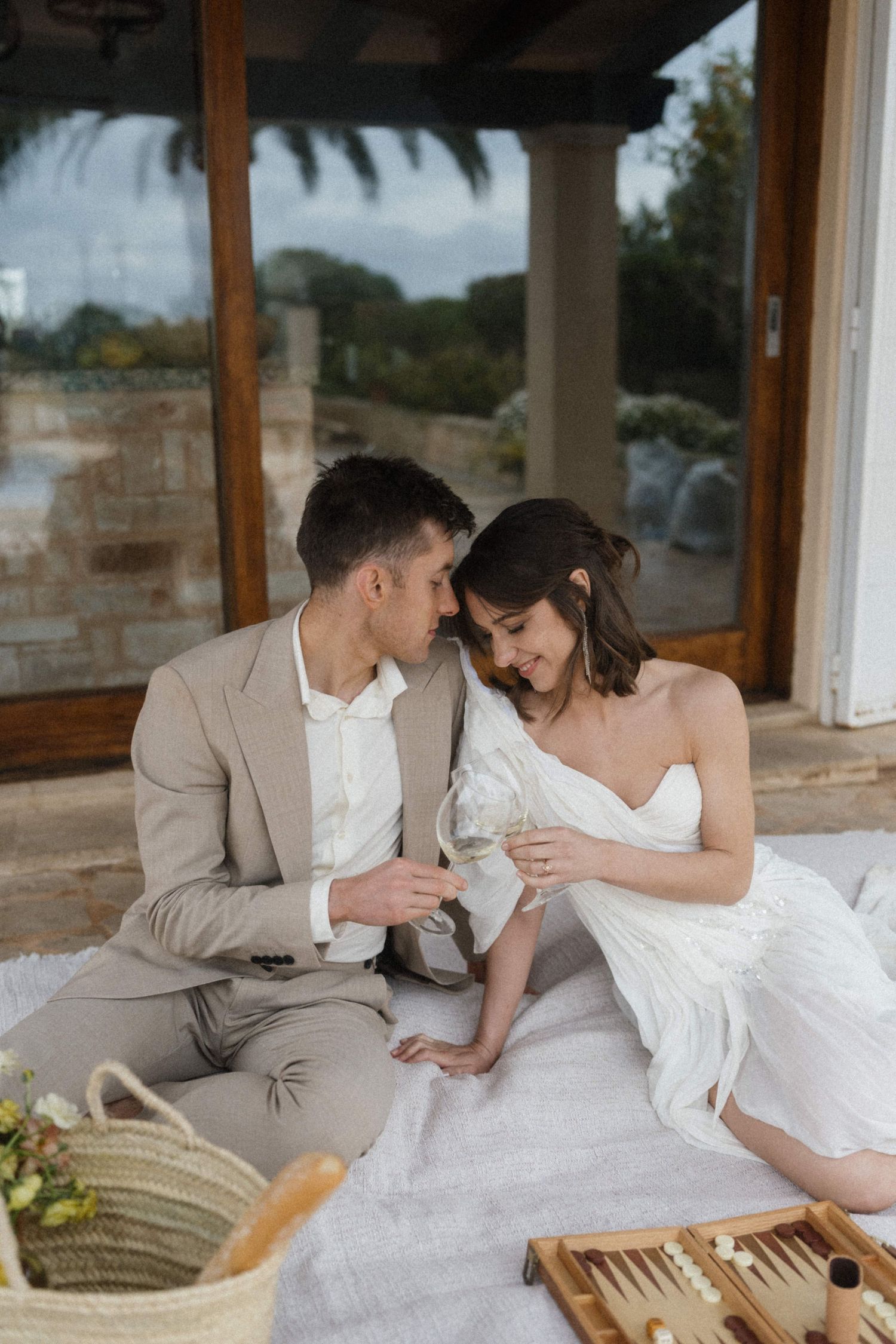 Elegant couple in wedding attire share an intimate moment during a romantic outdoor picnic with champagne.