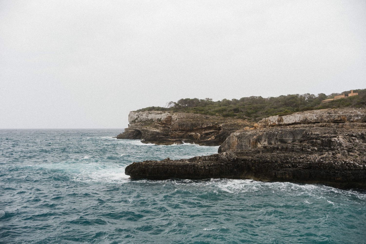 Rocky coastal cliffs overlooking turquoise ocean waves under an overcast sky in a scenic Mediterranean landscape.