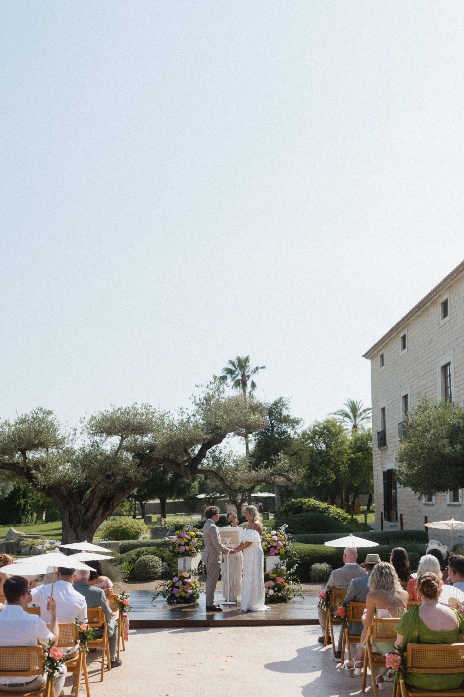 Outdoor wedding ceremony with couple at altar, guests seated in wooden chairs under white parasols.