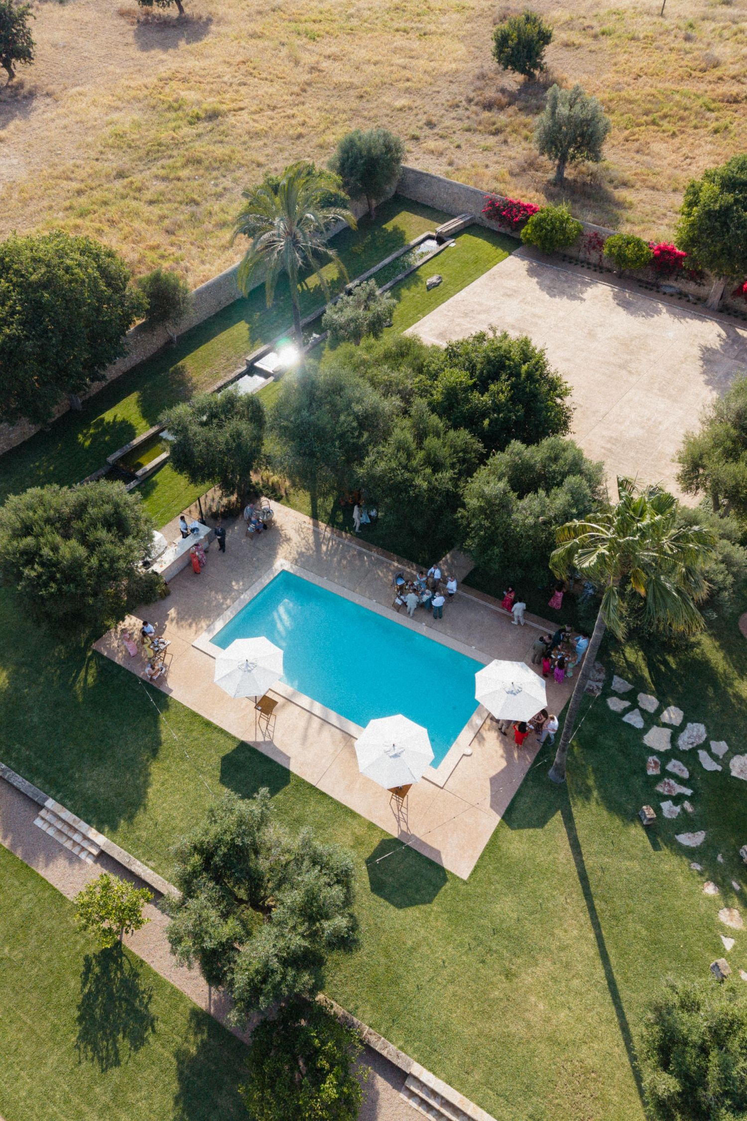 Aerial drone view of a rectangular swimming pool surrounded by wedding guests and white umbrellas.