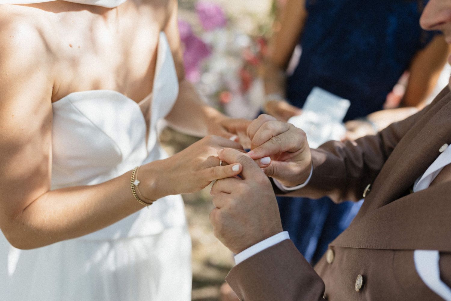 Bride placing a wedding ring on groom's finger during an outdoor ceremony, wearing a white strapless dress.