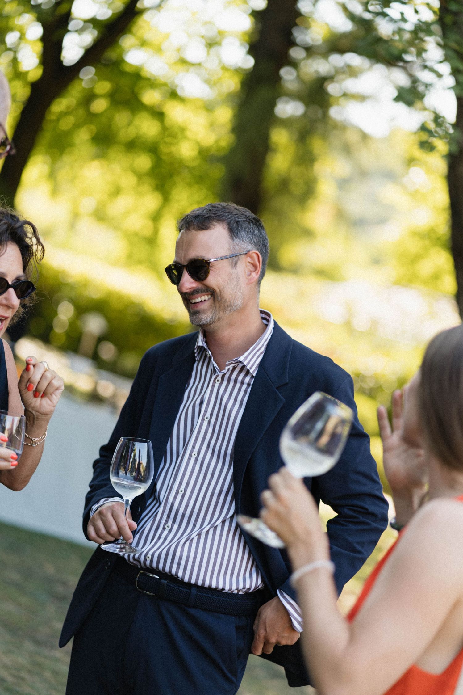 Smiling man in striped shirt and sunglasses enjoying wine at an outdoor garden party.