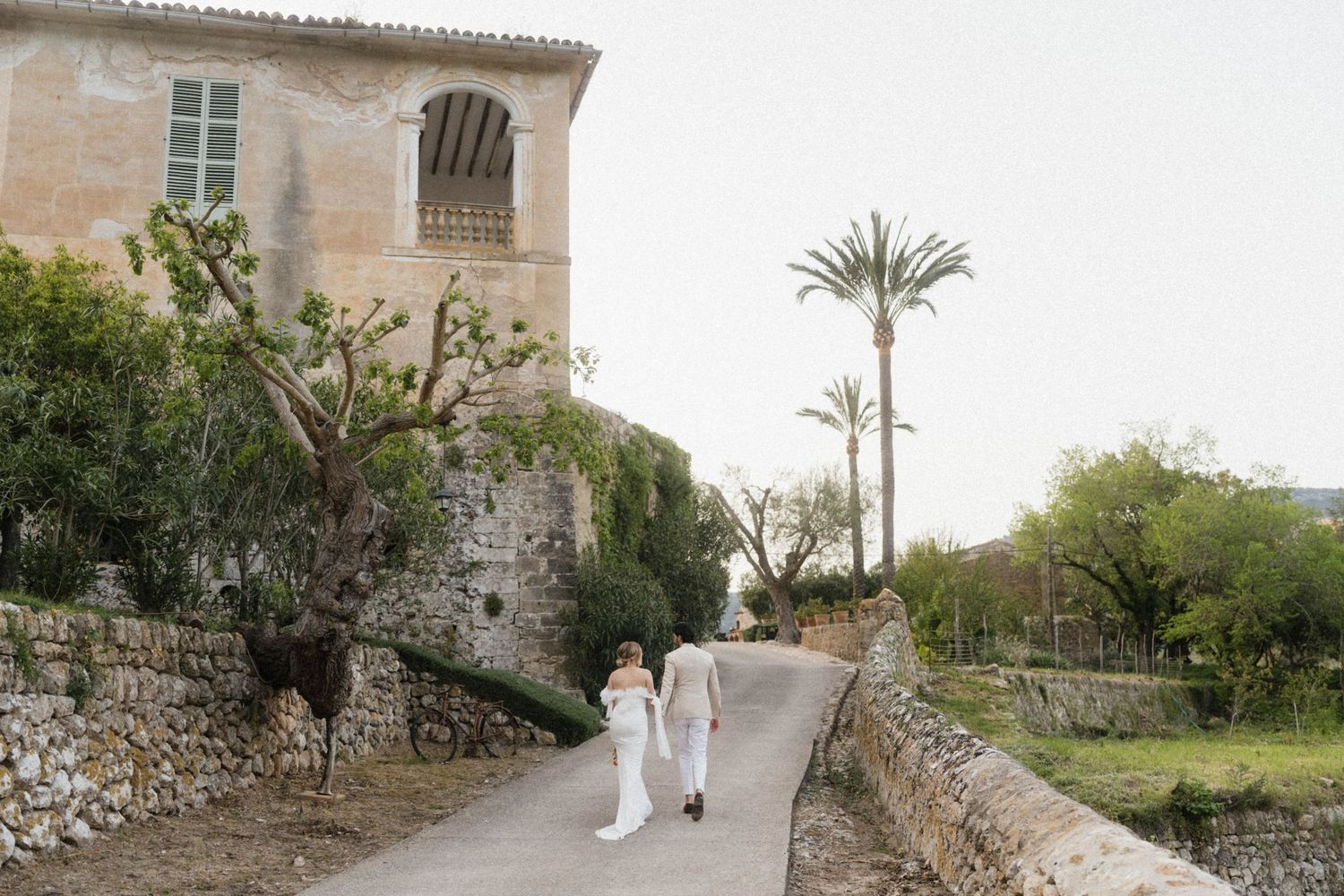 Bride and groom walk hand in hand along a stone path beside a historic building with palm trees.