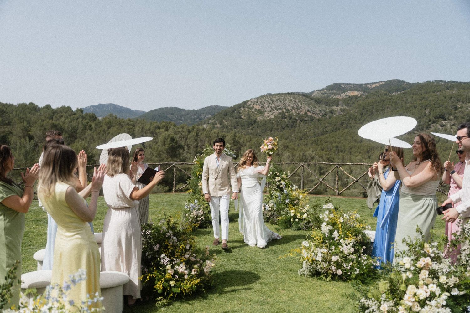 Bride and groom celebrate at outdoor wedding ceremony on green lawn with mountain views and white parasols.