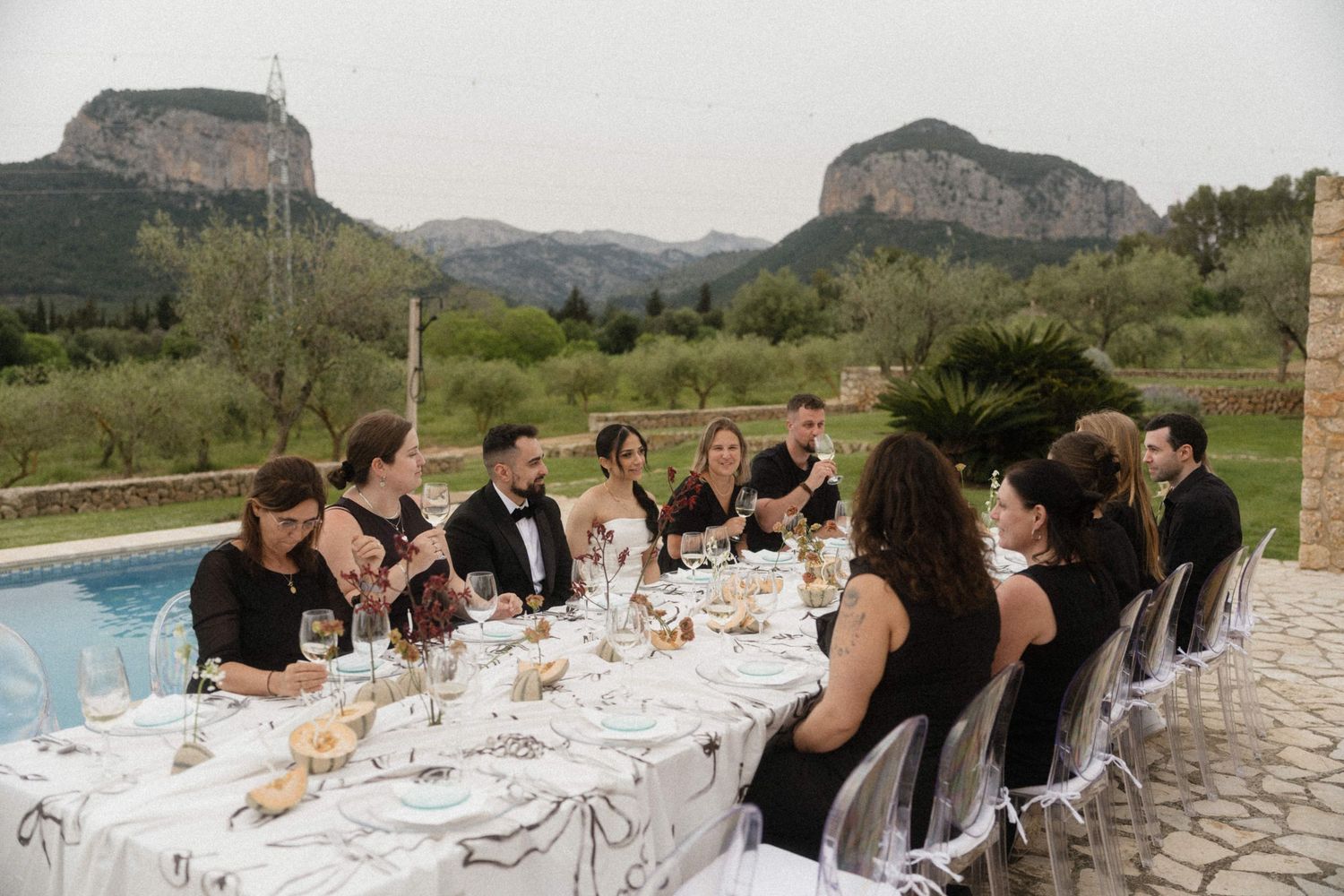 Wedding guests dining al fresco at elegant table beside pool with dramatic mountain backdrop.