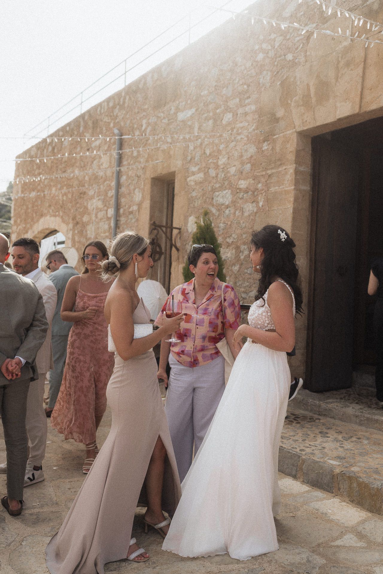 Bride in white gown chats with guests outside a rustic stone venue on a sunny wedding day.