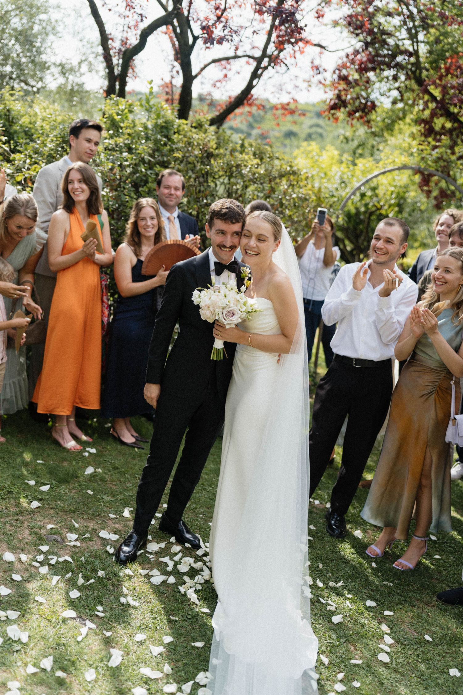 Joyful newlyweds celebrate with guests tossing white rose petals in a sunlit garden after their Italian wedding ceremony.