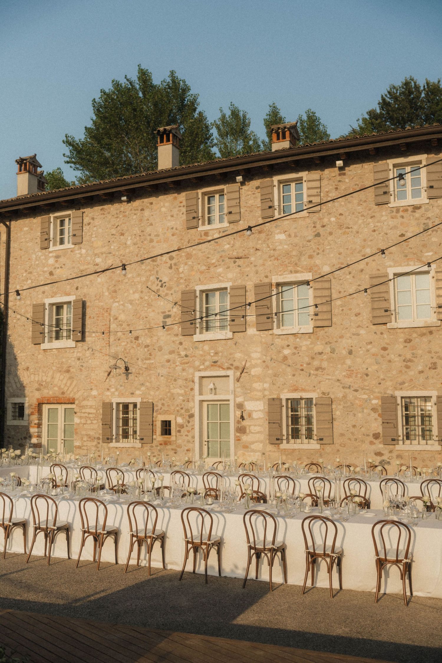 Long outdoor wedding reception tables set before a rustic Italian stone villa with string lights at dusk.