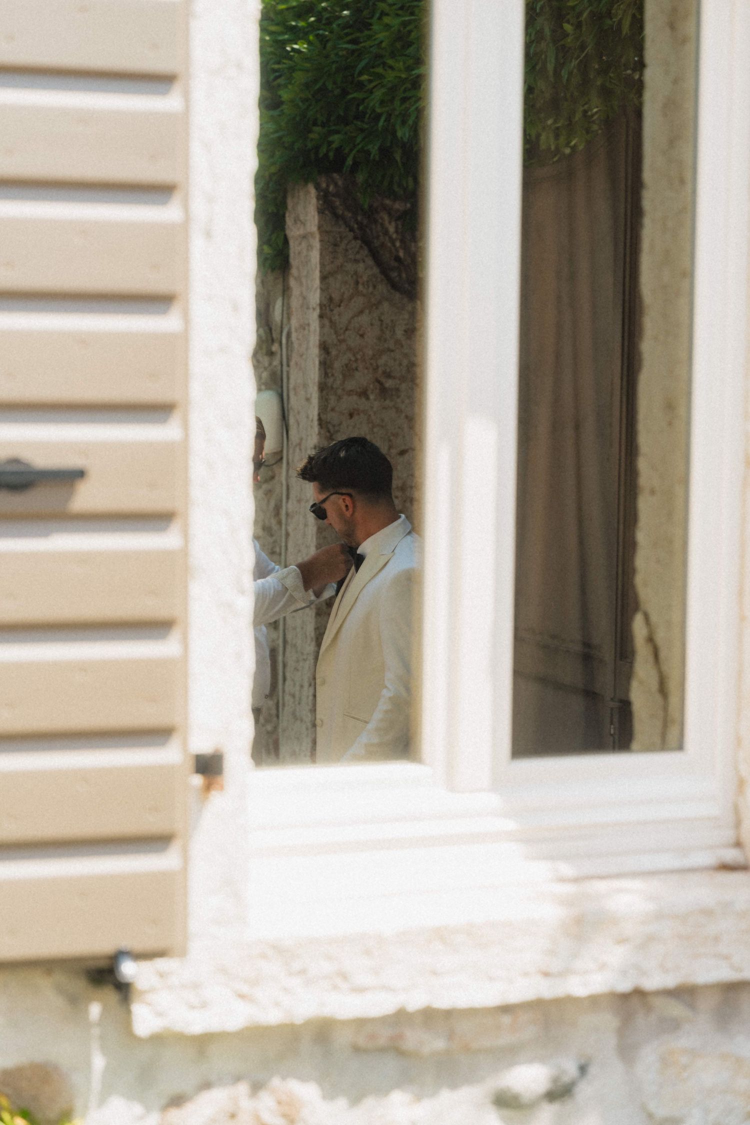 Man in white suit and sunglasses standing in a doorway framed by white shutters and lush greenery.
