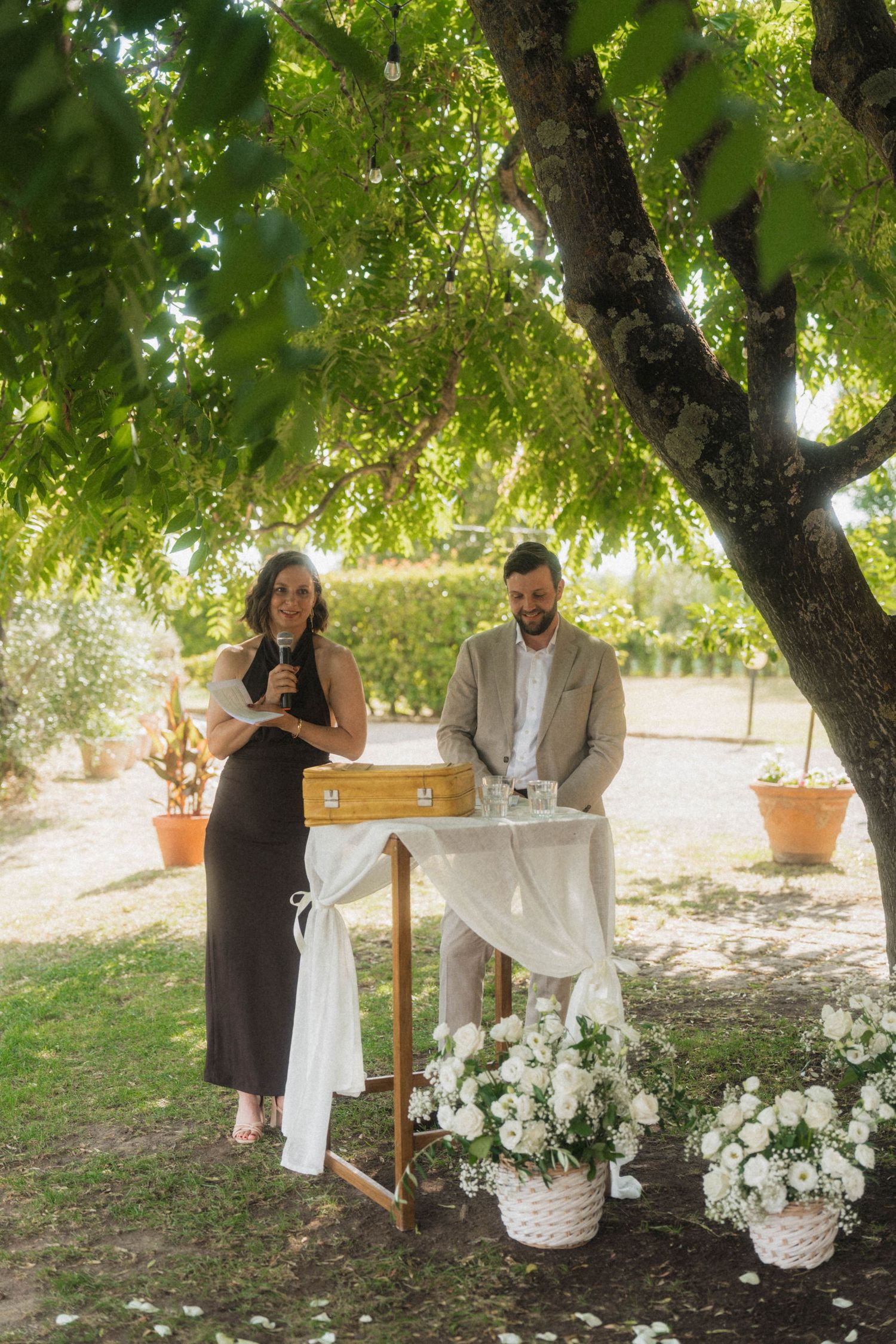Officiant and groom stand at a decorated outdoor wedding ceremony altar beneath a sunlit tree.