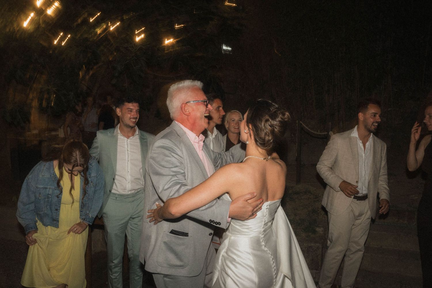 Bride sharing a dance with an older gentleman at an outdoor evening wedding reception under warm string lights.