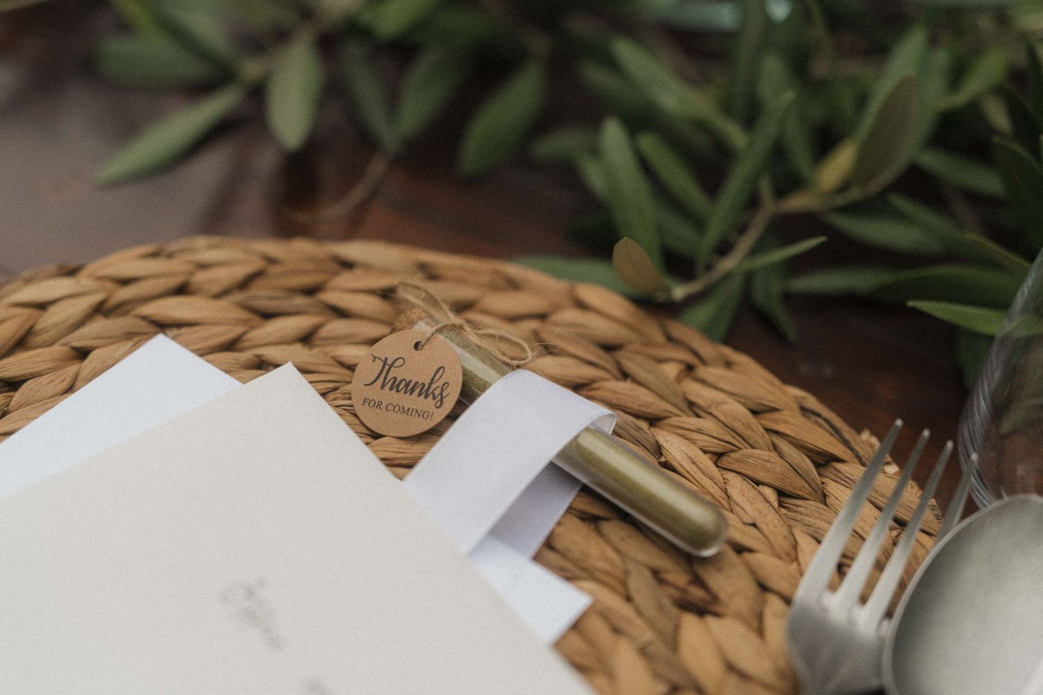 Elegant wedding place setting with woven placemat, white menu card, and wooden thank you tag on table.
