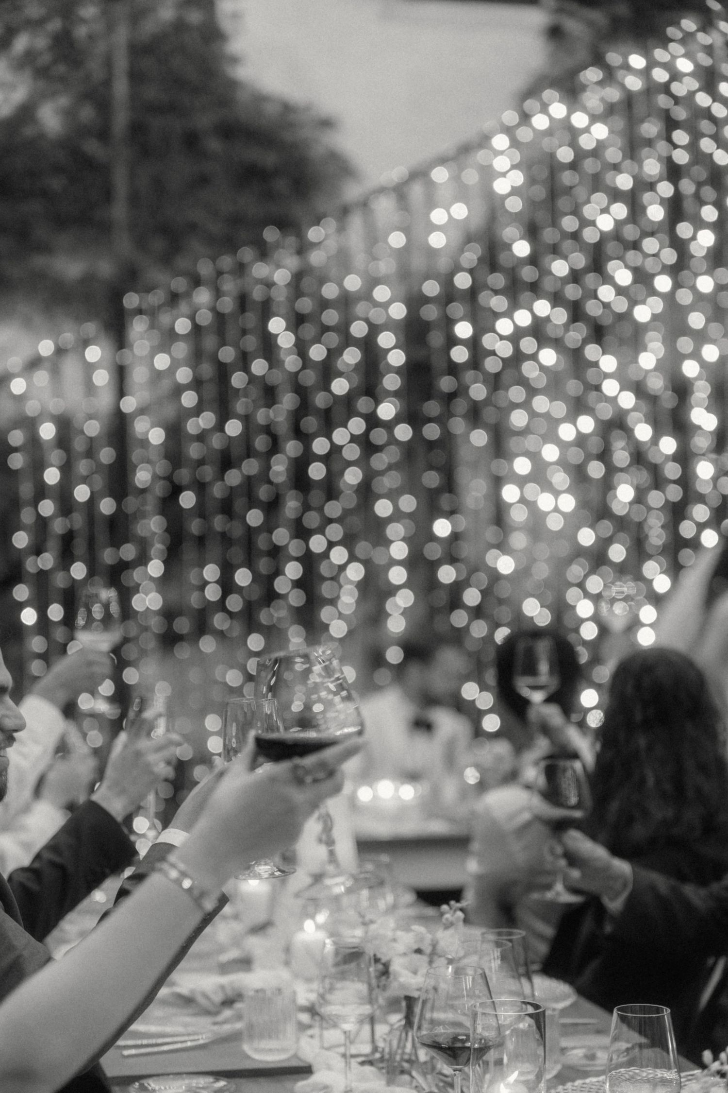 Guests raising wine glasses in toast at outdoor wedding reception with bokeh lights.
