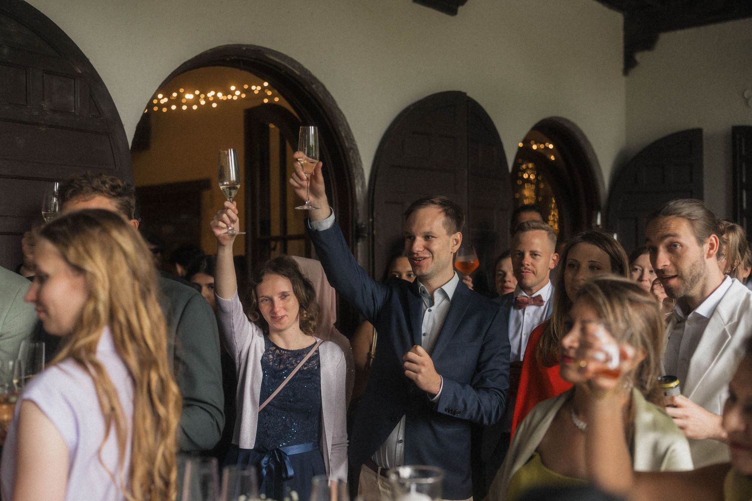 Wedding guests raising champagne glasses in a toast inside a rustic venue with arched doorways.