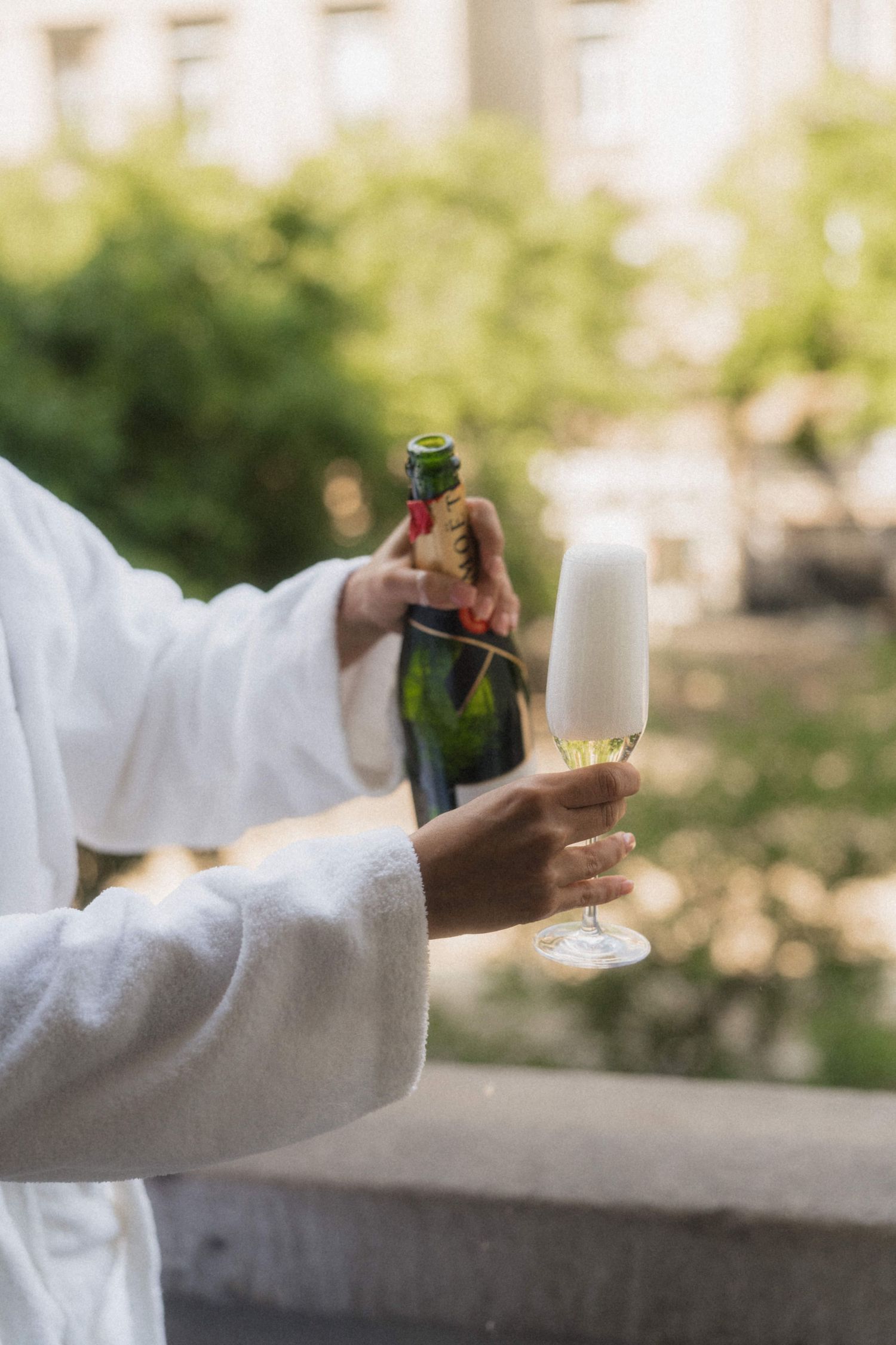 Person in white robe pouring champagne into a flute glass on a balcony with green trees in background.