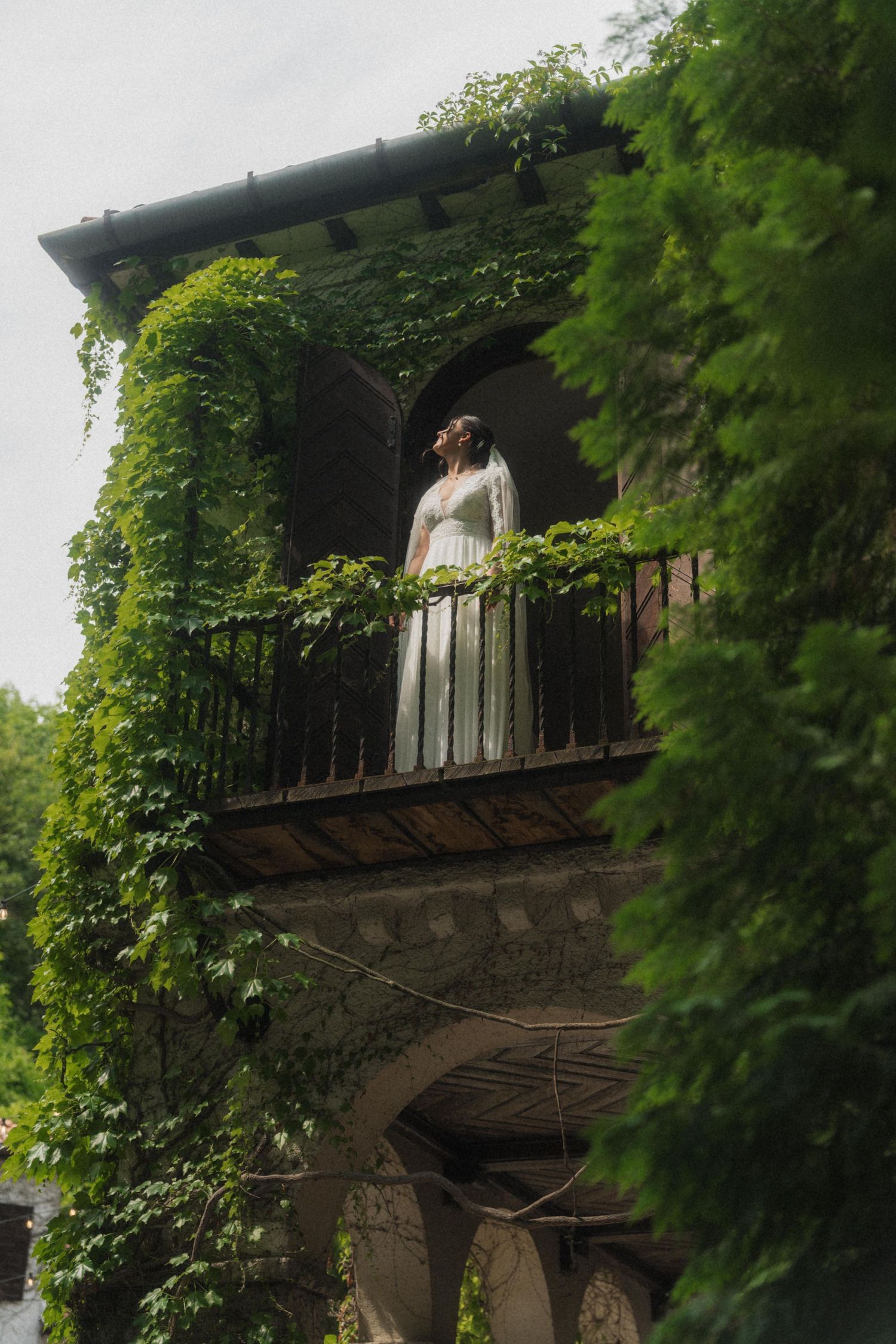Bride in white gown standing on ivy-covered stone balcony, gazing upward at overcast sky.