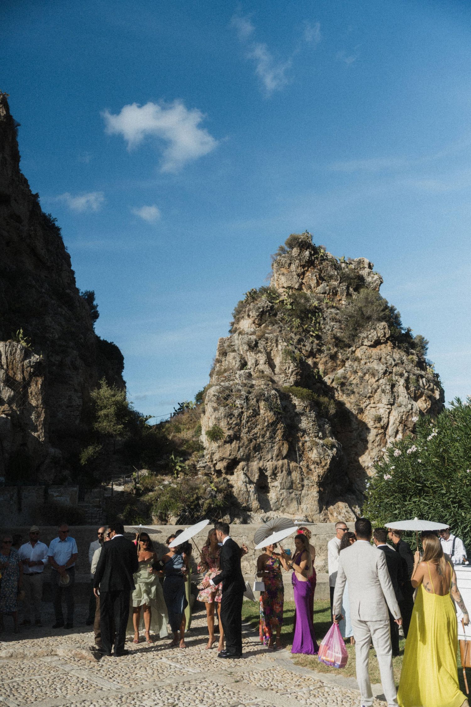 Wedding guests gathered outdoors near dramatic rocky cliffs under a bright blue sky.
