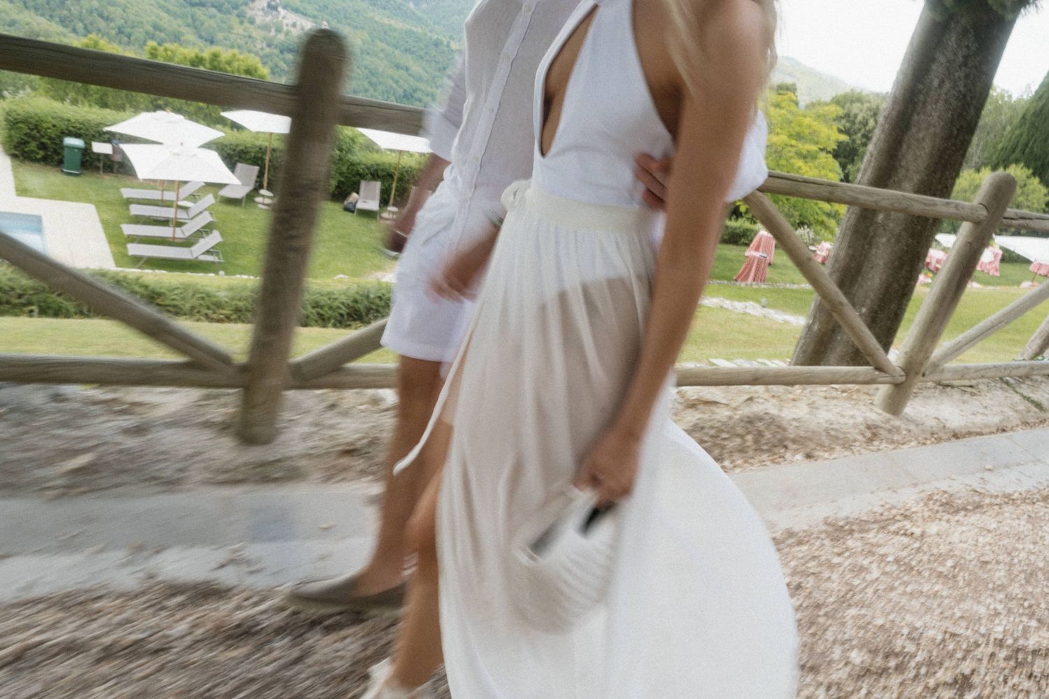 Couple in white outfits walking along a wooden fence path at an outdoor wedding venue.