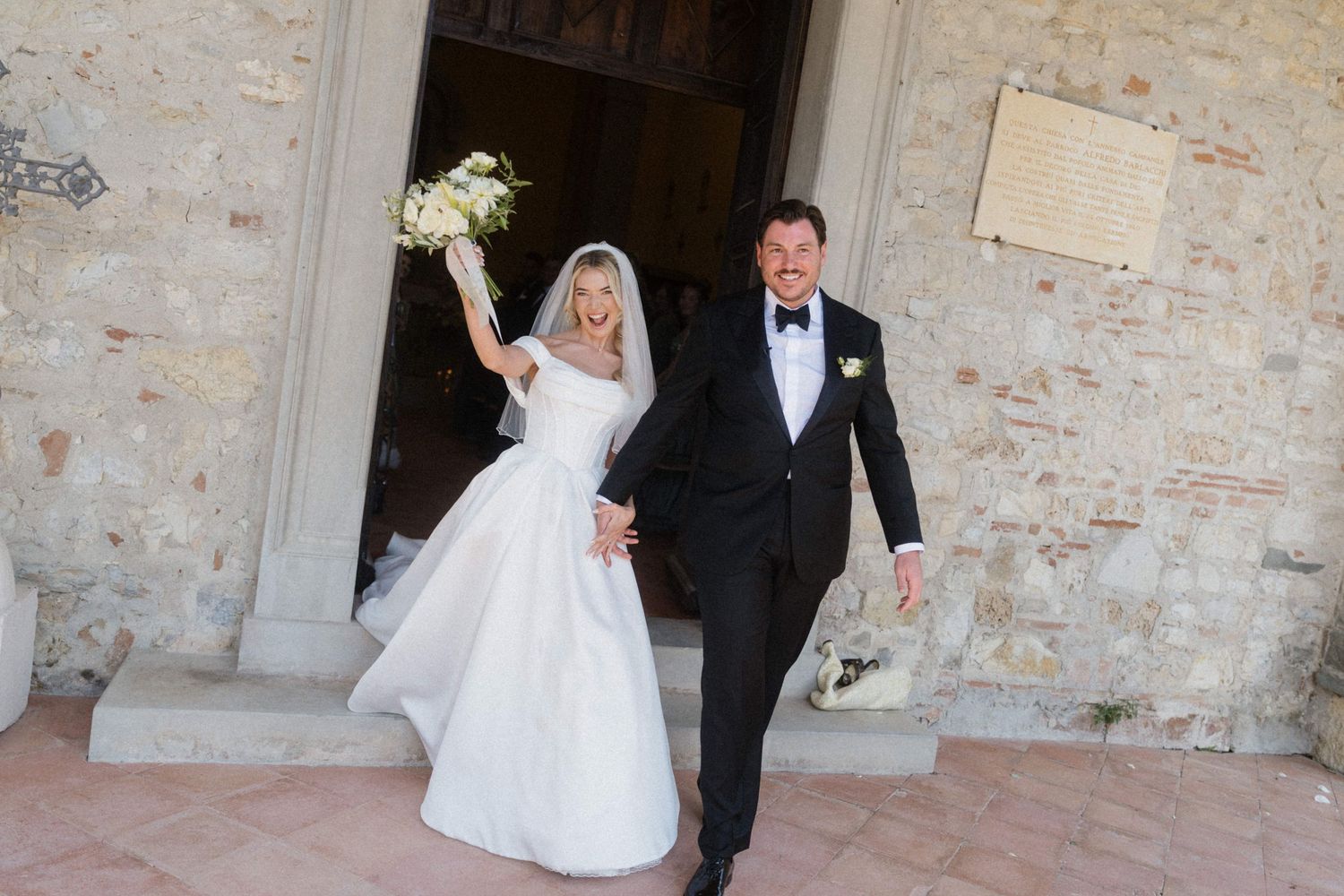 Joyful bride and groom exit historic stone church, bride raising bouquet of white flowers in celebration.