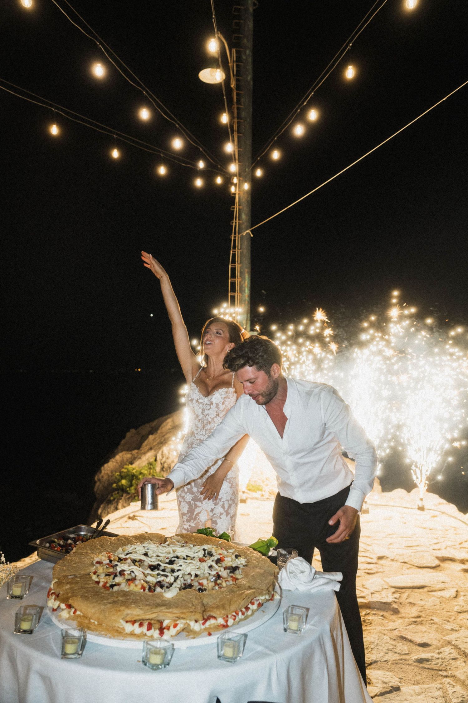 Bride and groom cut large wedding pizza cake outdoors at night with fireworks behind them.