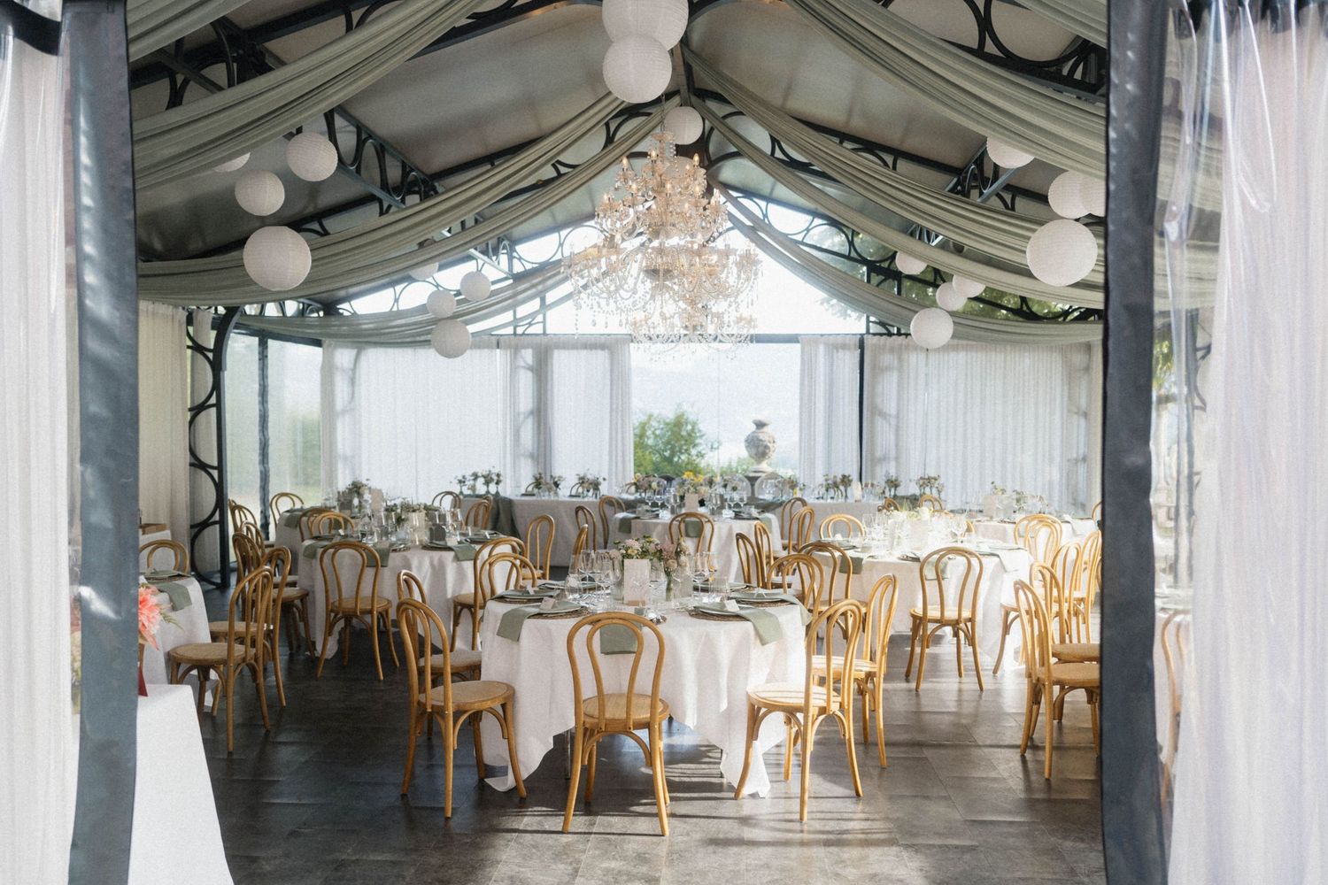 Elegant wedding reception tent with chandelier, round tables, and gold Thonet chairs.