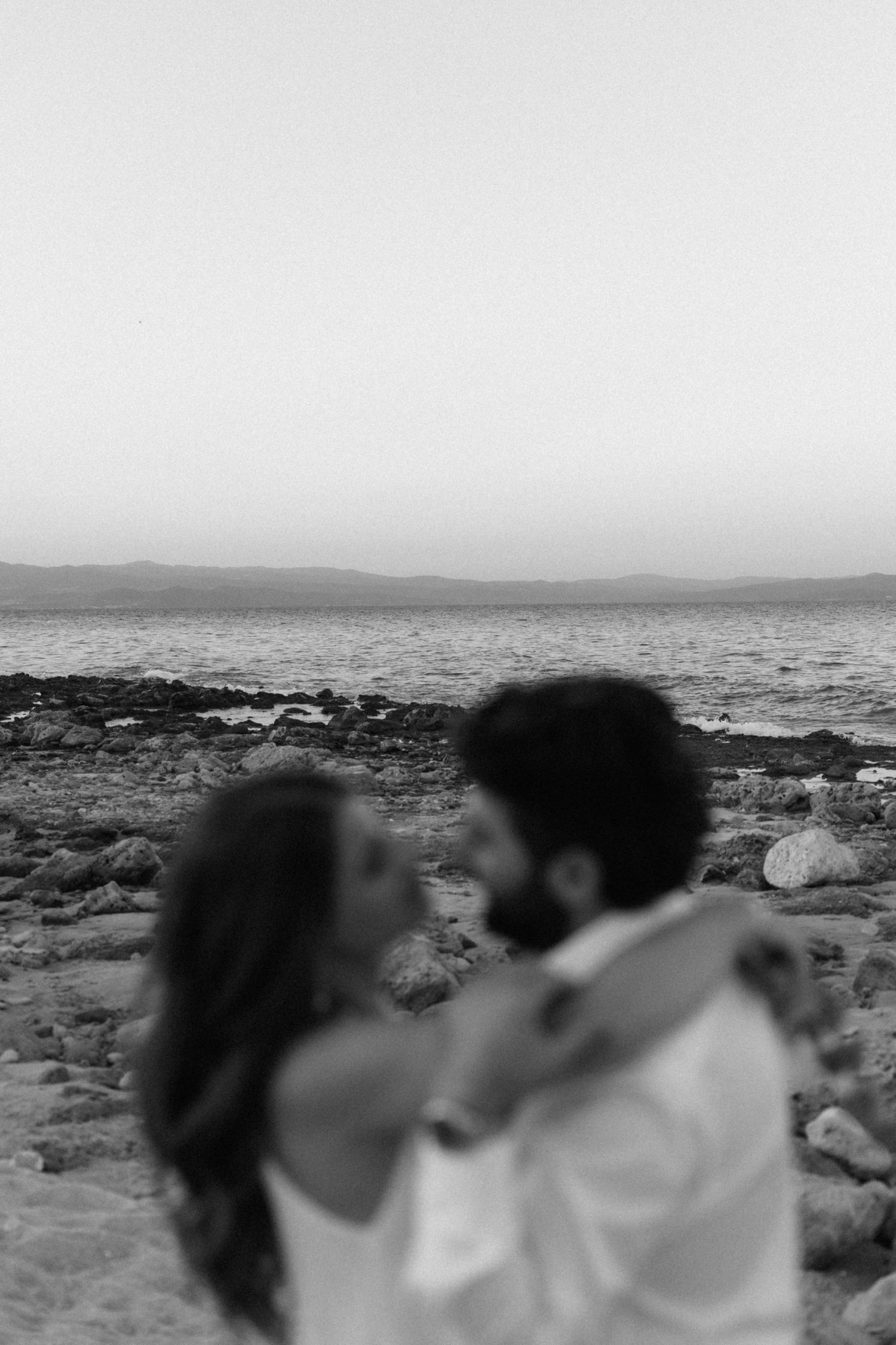 Black and white blurred romantic photo of couple about to kiss on rocky shoreline at dusk.