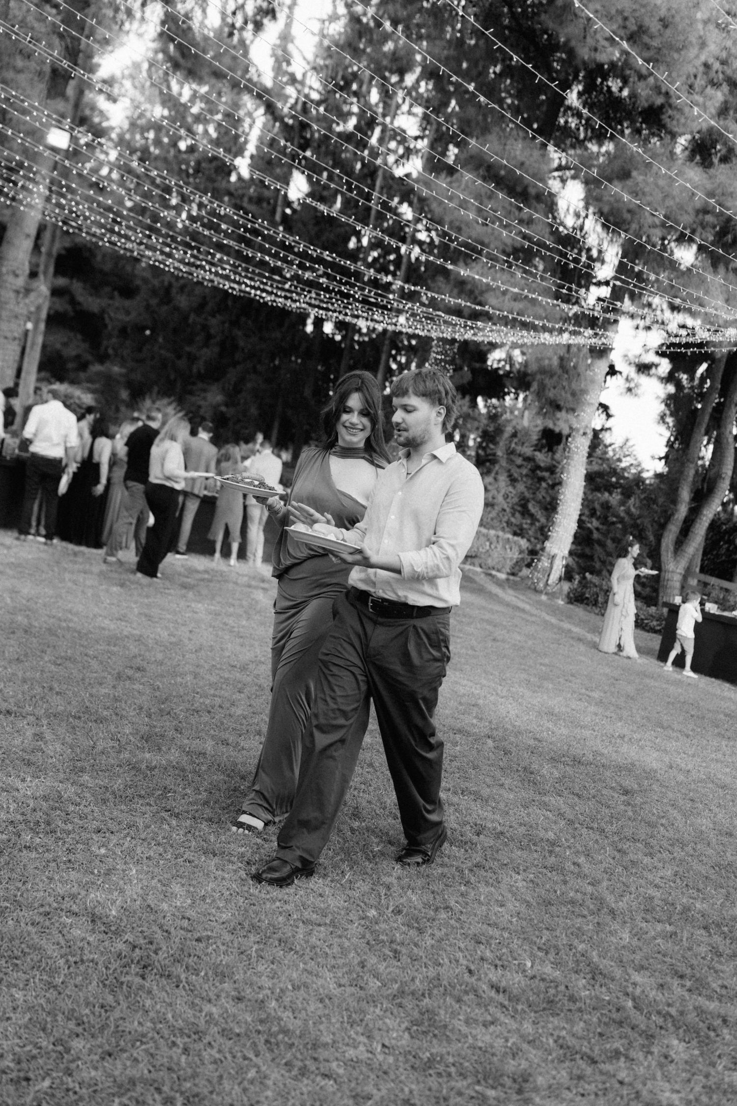 Couple dancing on grass under string lights at outdoor wedding reception in black and white.