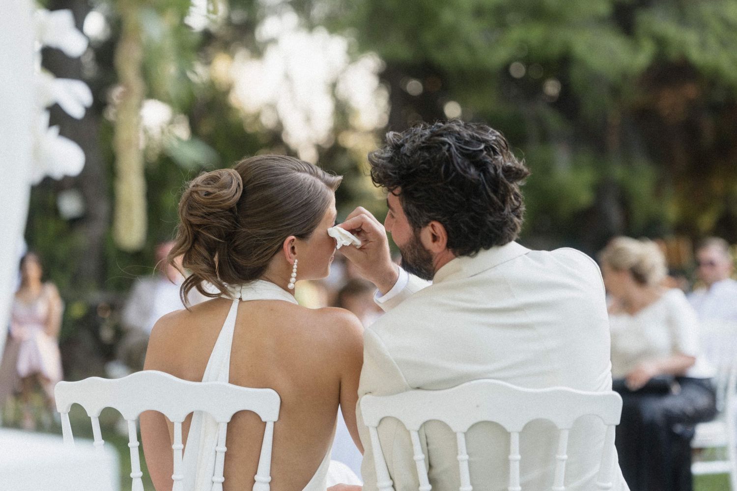 Groom tenderly touches brides face while seated at outdoor wedding ceremony surrounded by guests.