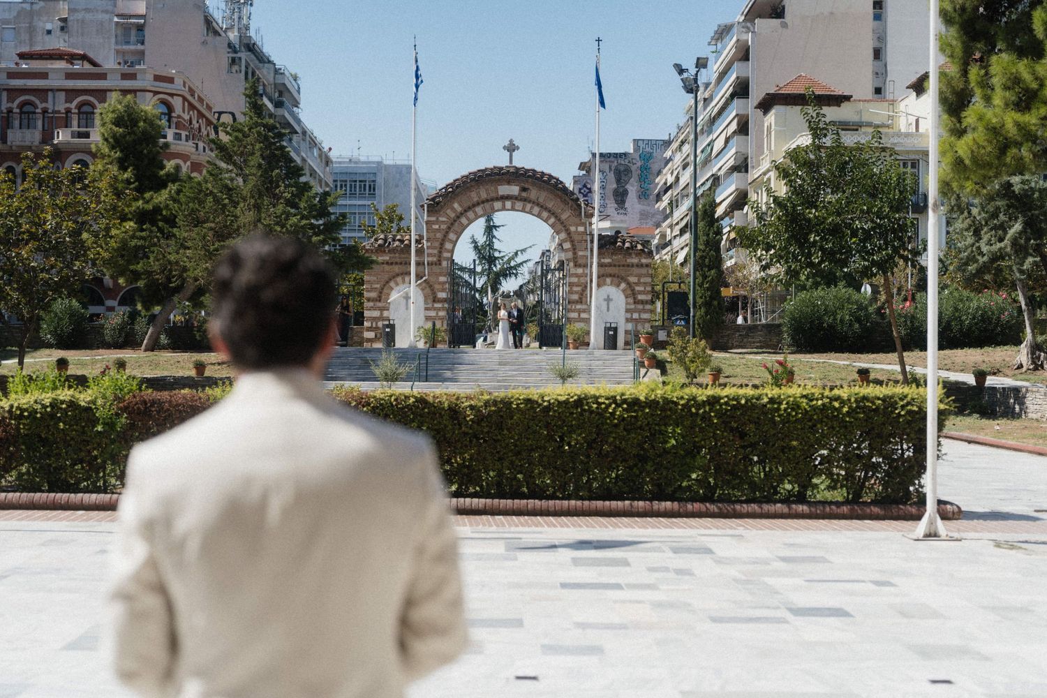 Person in beige jacket views ornate arch monument in a sunny urban plaza surrounded by greenery.