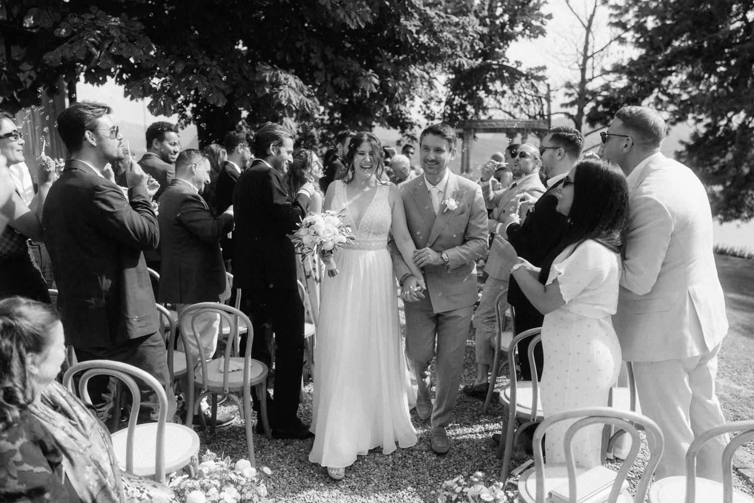 Black and white photo of newlyweds walking back down aisle as guests applaud at outdoor wedding.