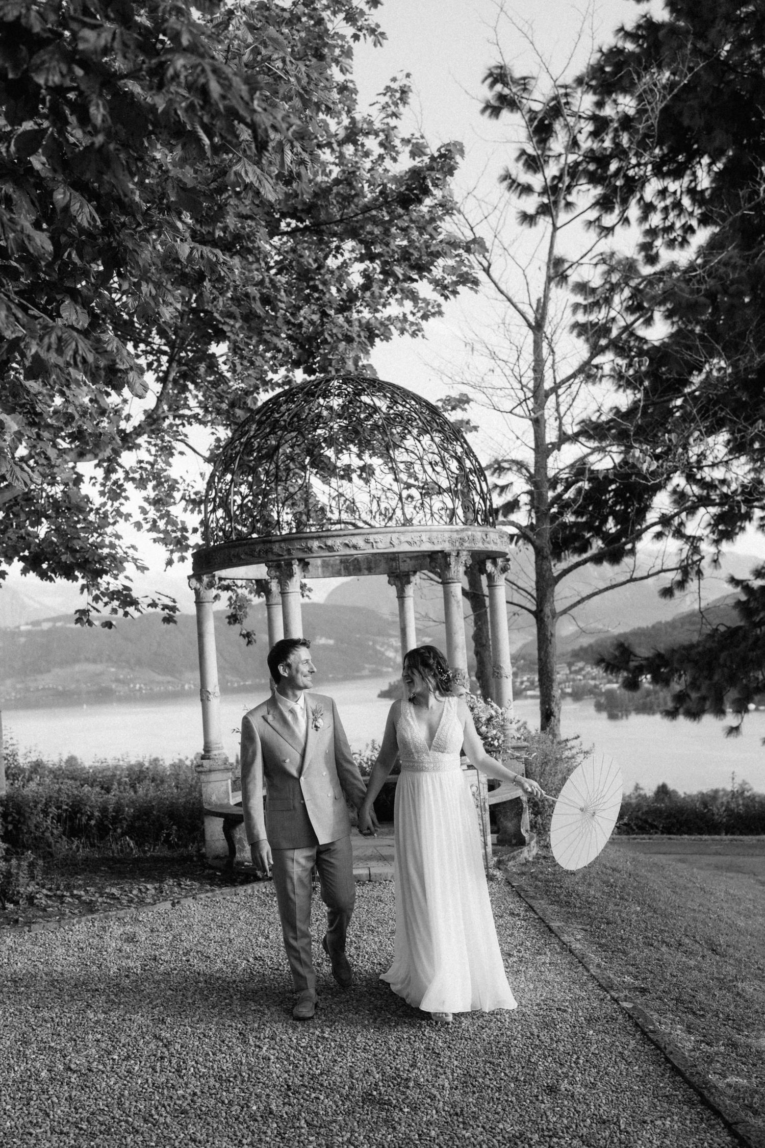 Bride and groom hold hands near ornate gazebo with lake and mountain backdrop in black and white.