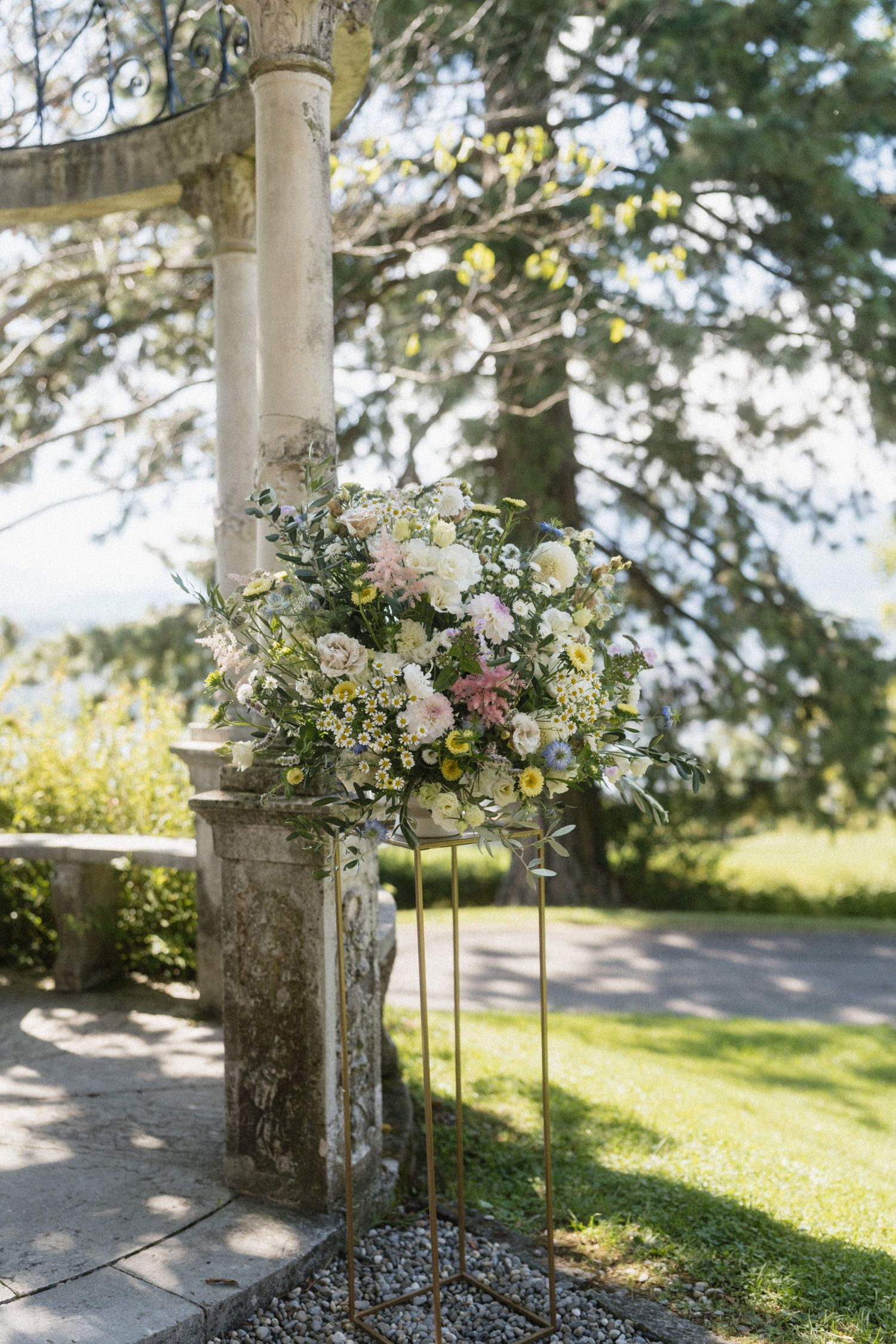 Elegant floral arrangement with pink and white blooms on a gold stand beside a stone column at an outdoor wedding.