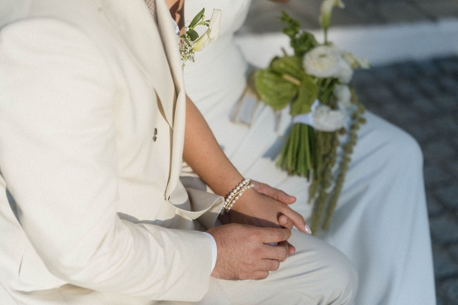 Close-up of wedding couple in white outfits, bride wearing pearl bracelet, groom in white suit with boutonniere holding hands.