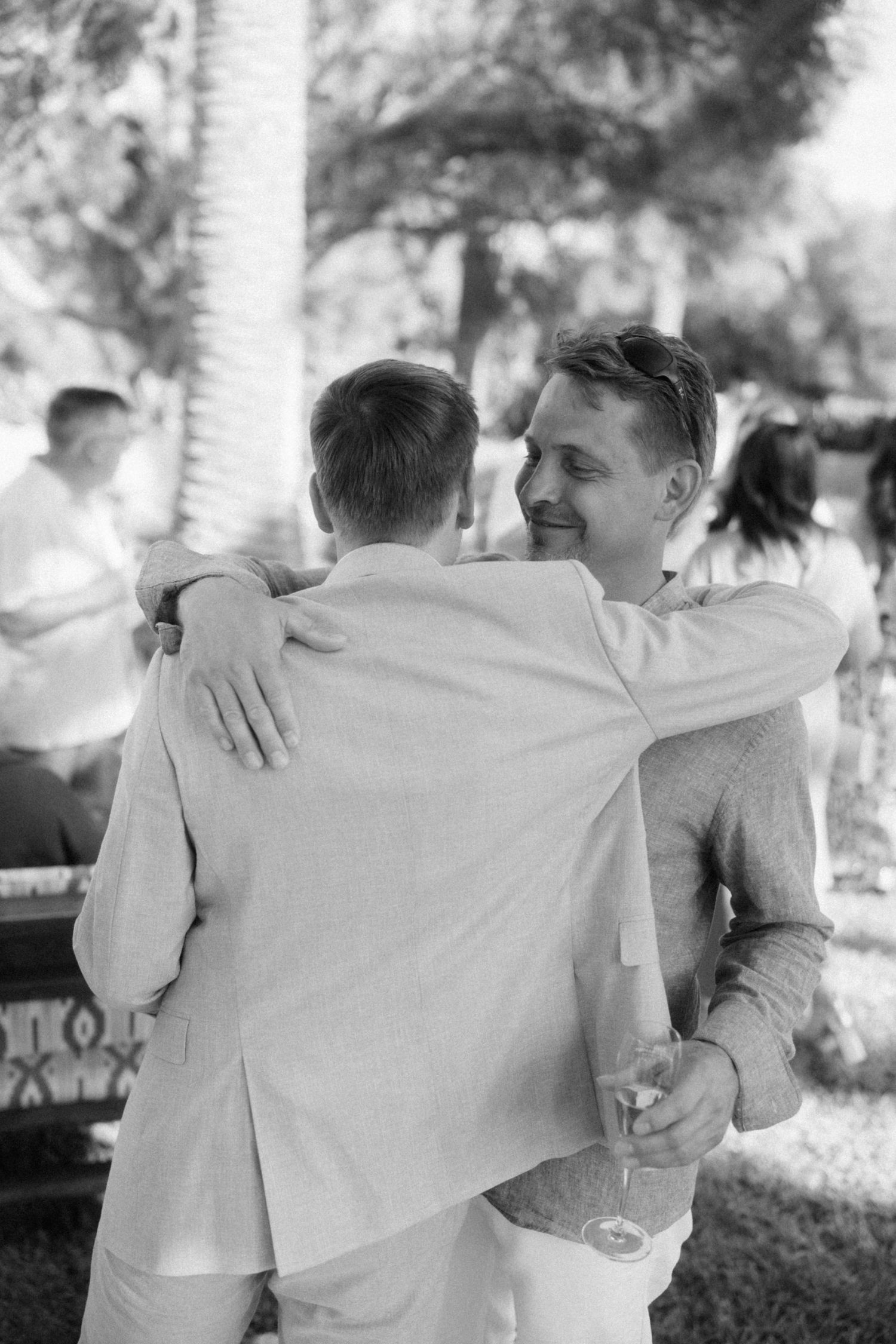 Two men share a warm embrace at an outdoor wedding reception in black and white photography.