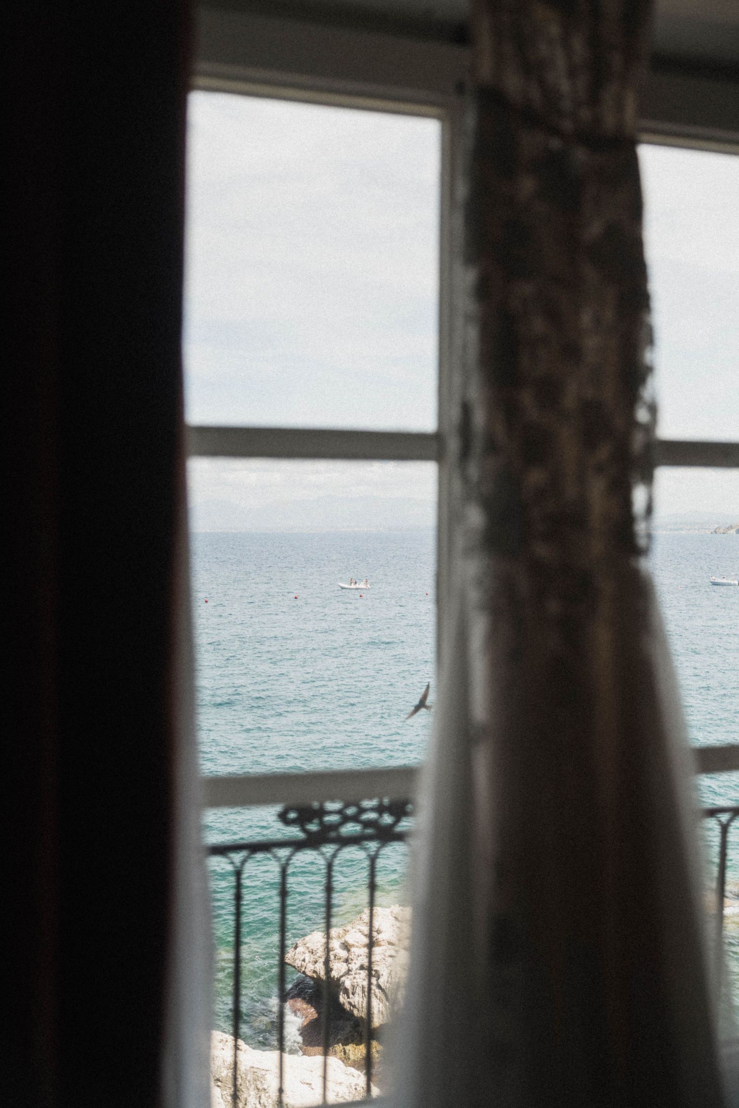 Elegant wedding dress hanging by window with breathtaking ocean view and iron balcony railing.