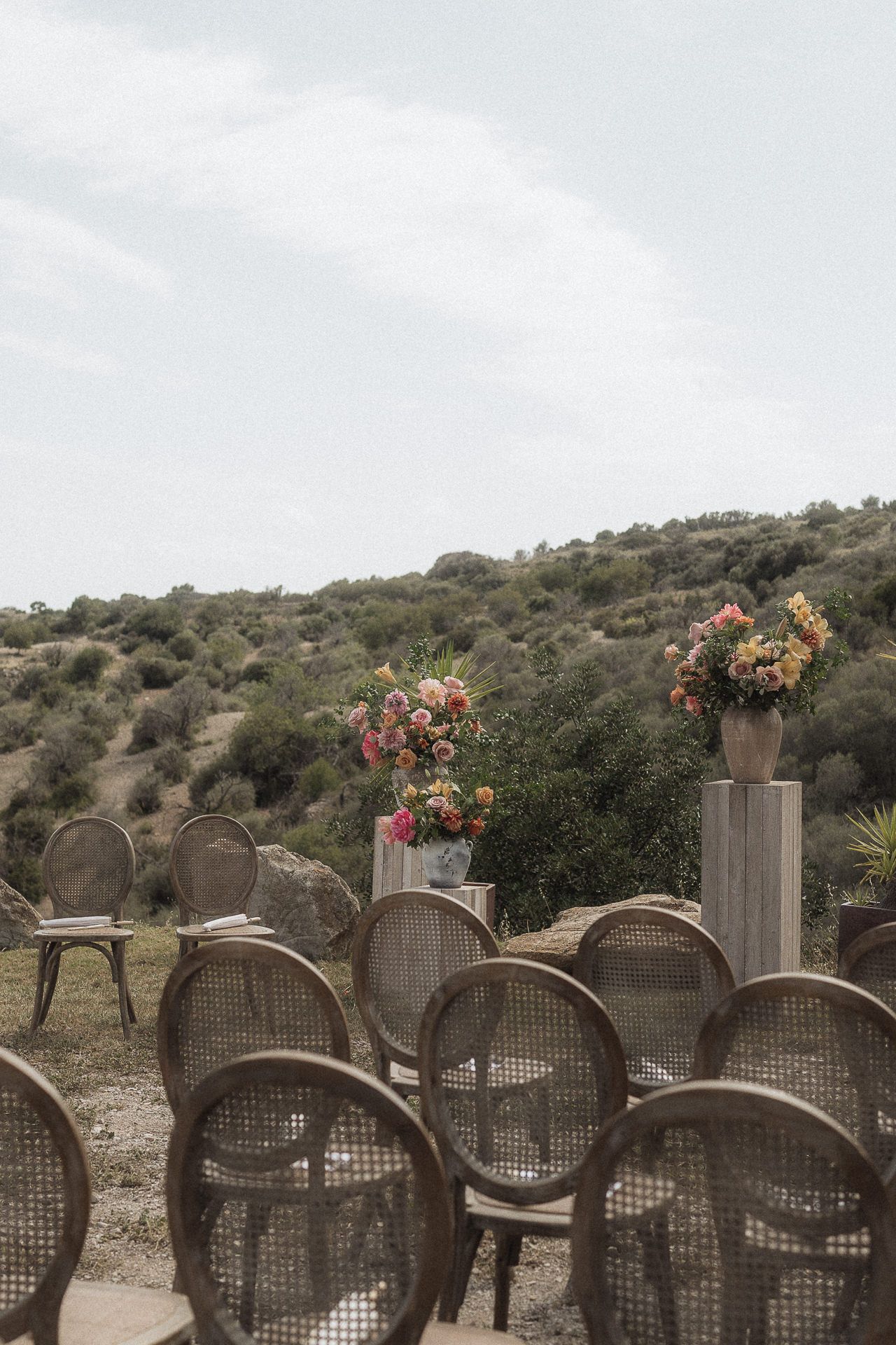 Rustic cane chairs arranged outdoors for a wedding ceremony with colorful floral arrangements on pedestals against a hillside backdrop.
