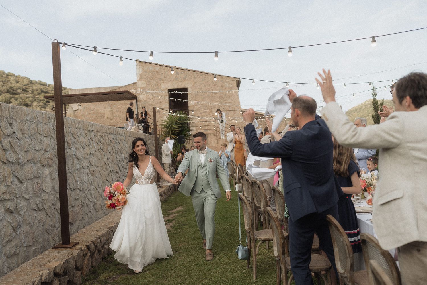 Bride and groom walk through cheering guests at an outdoor rustic wedding reception with string lights.