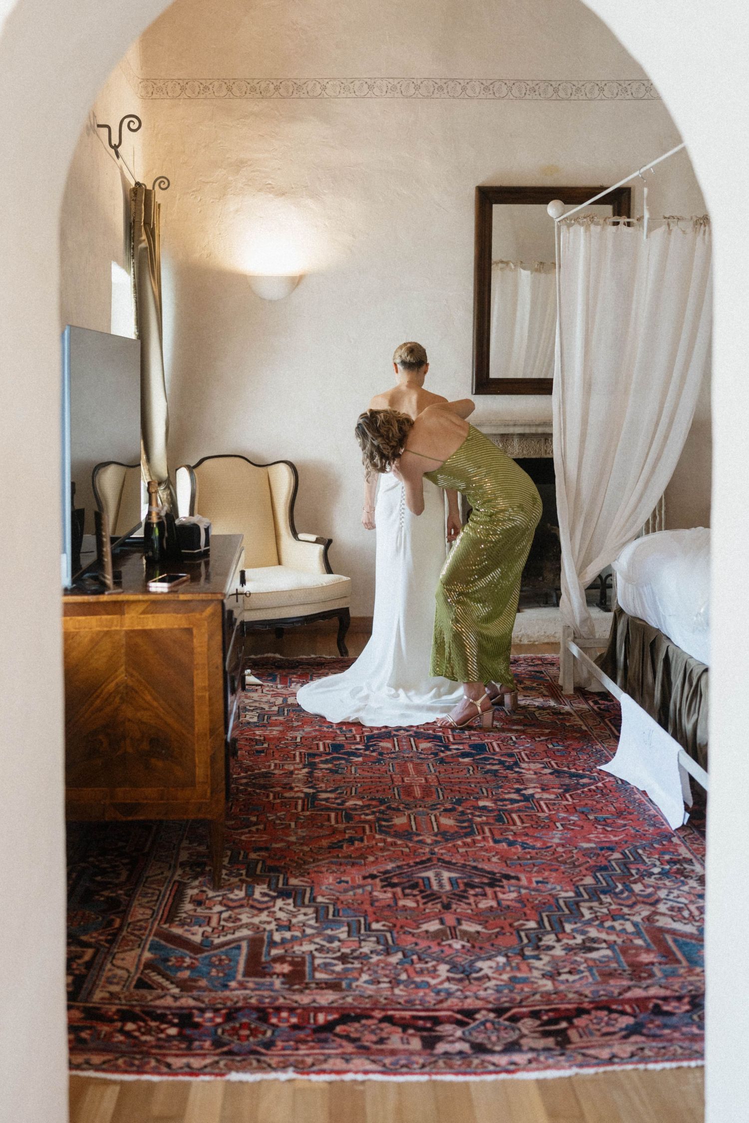 Bridesmaid in green dress helps bride into white wedding gown on ornate red Persian rug in elegant room.