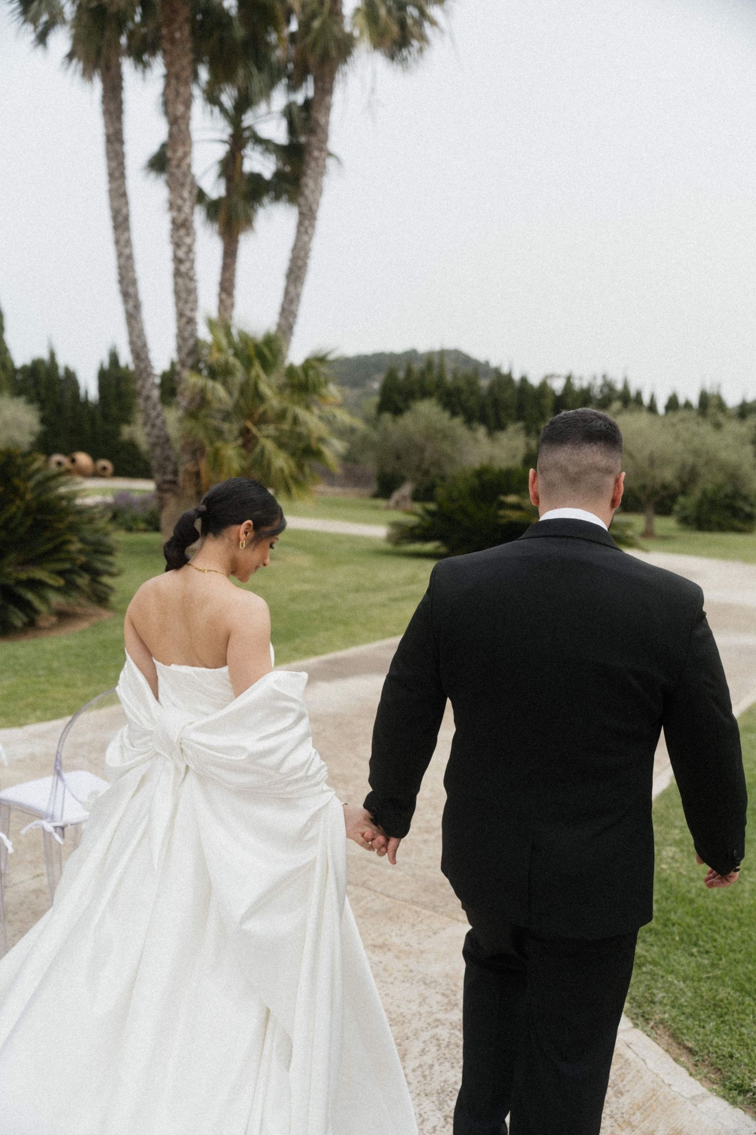 Bride in strapless white gown with bow and groom in black tuxedo walk hand in hand through lush garden.