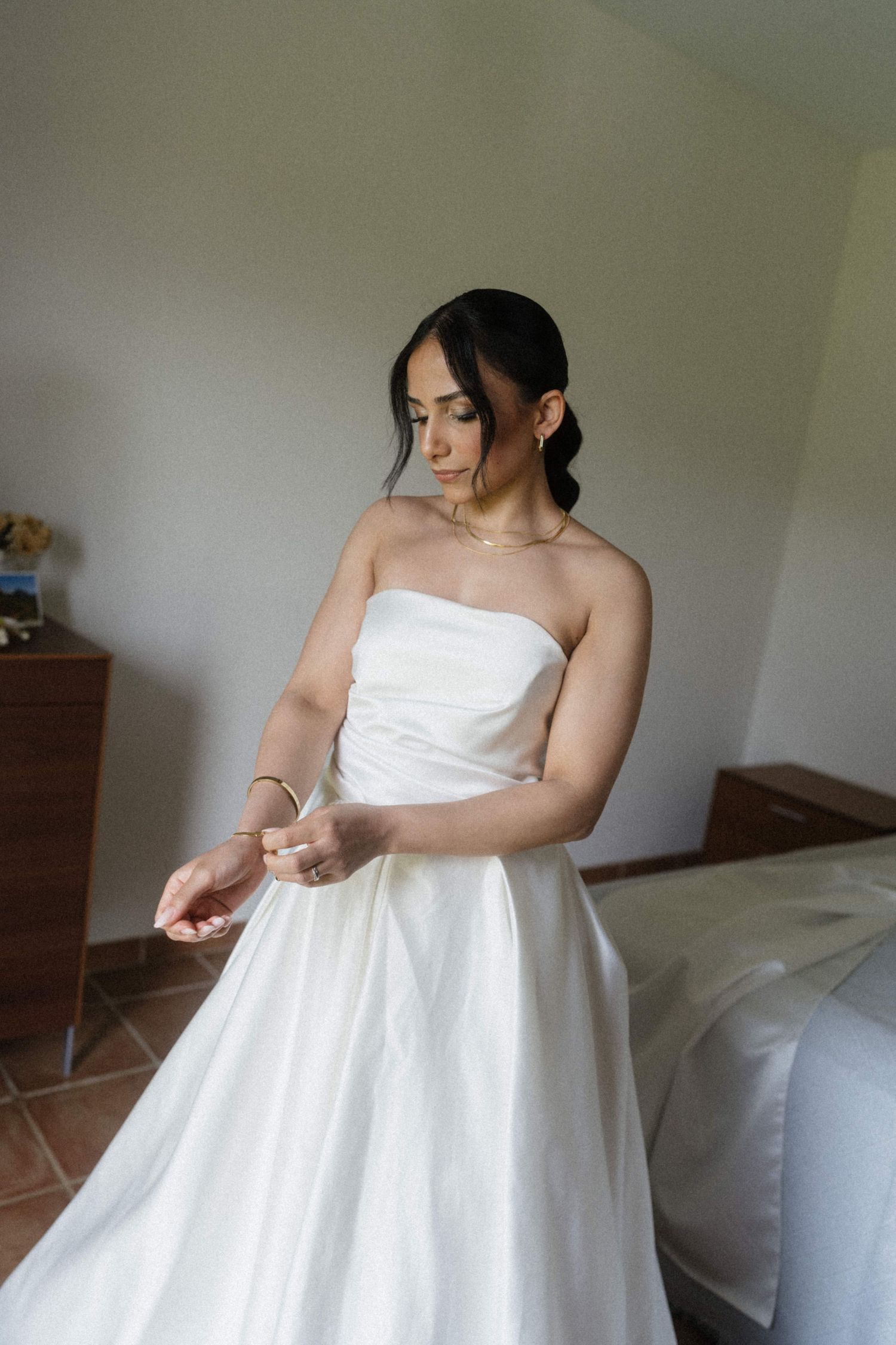 Bride in strapless white ball gown adjusting her bracelet in a softly lit bedroom.