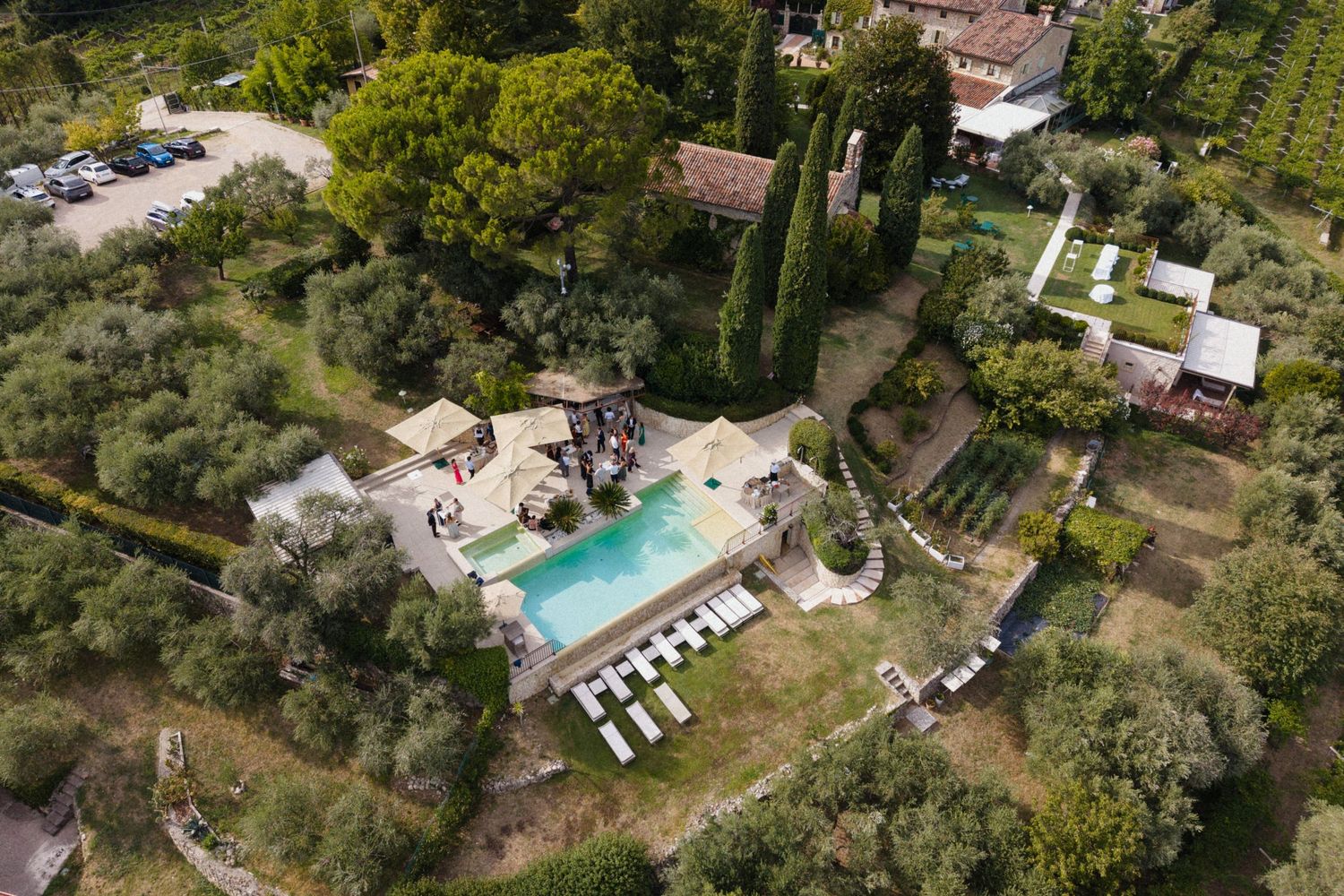 Aerial view of a Tuscan villa with a turquoise pool, surrounded by cypress trees, olive groves, and lush gardens.
