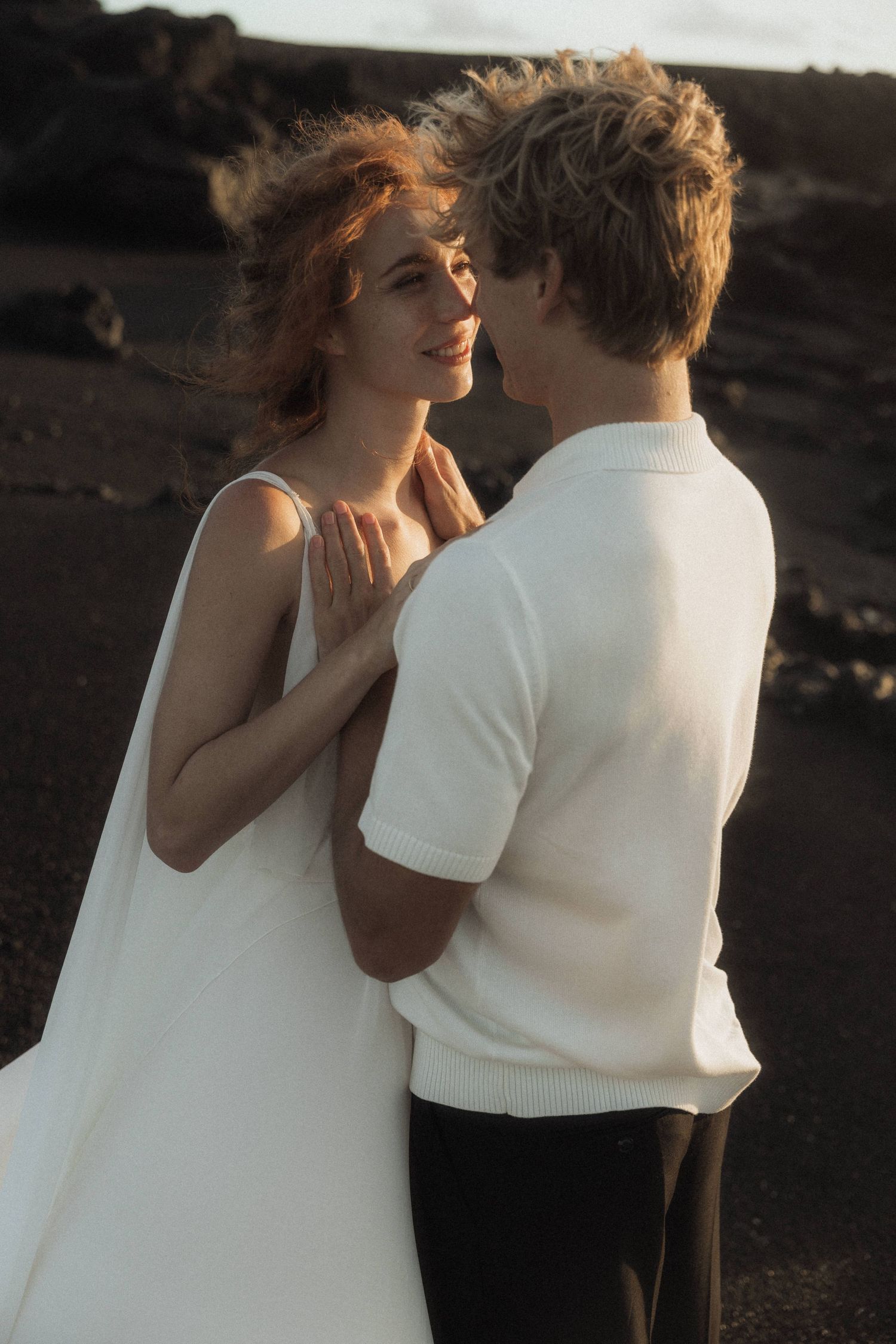 Bride smiles tenderly at groom on dark volcanic beach in warm sunset light.